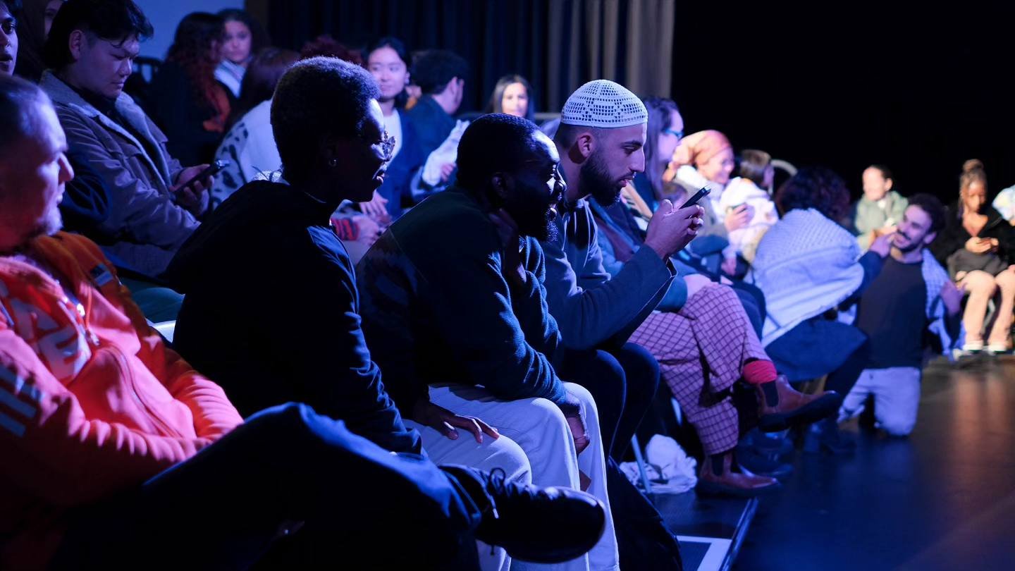 Audience sitting in dimly lit theater, some looking at their phones, engaged and waiting for a performance or event.
