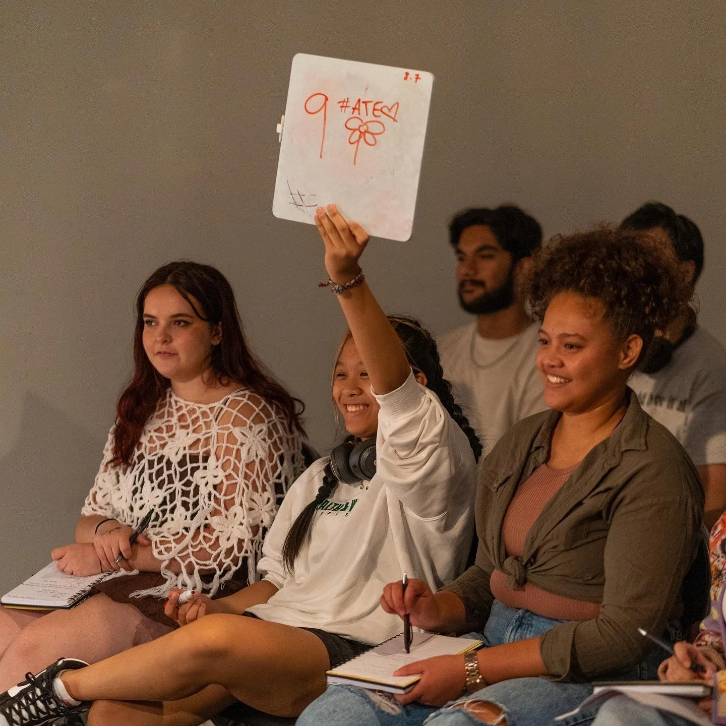 People sitting in an audience at an event, one woman raising a whiteboard with #ATEA written on it and a flower drawing, others taking notes and looking on.
