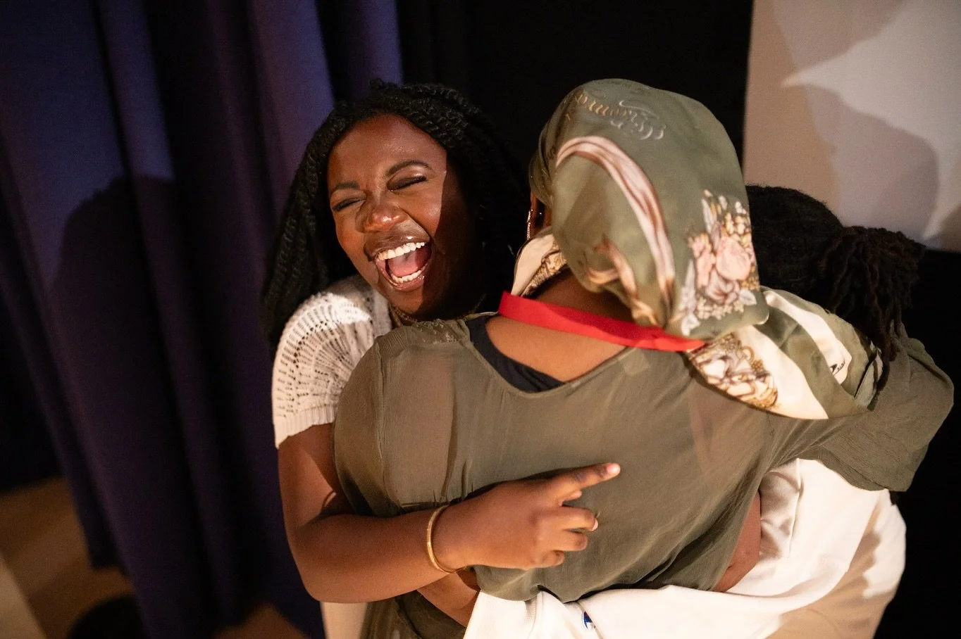 Two women hugging and laughing joyfully indoors, one woman with face visible and the other with head turned away, wearing a headscarf and casual clothing.