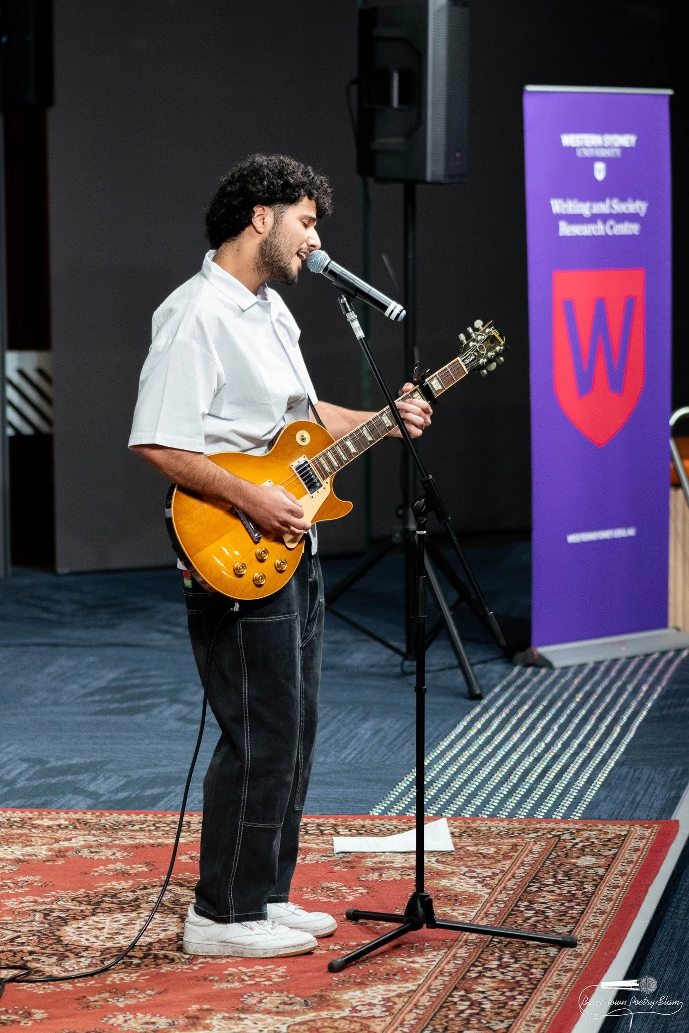 A young man is performing with an electric guitar and singing into a microphone on stage, with a purple banner for Western Sydney University in the background.