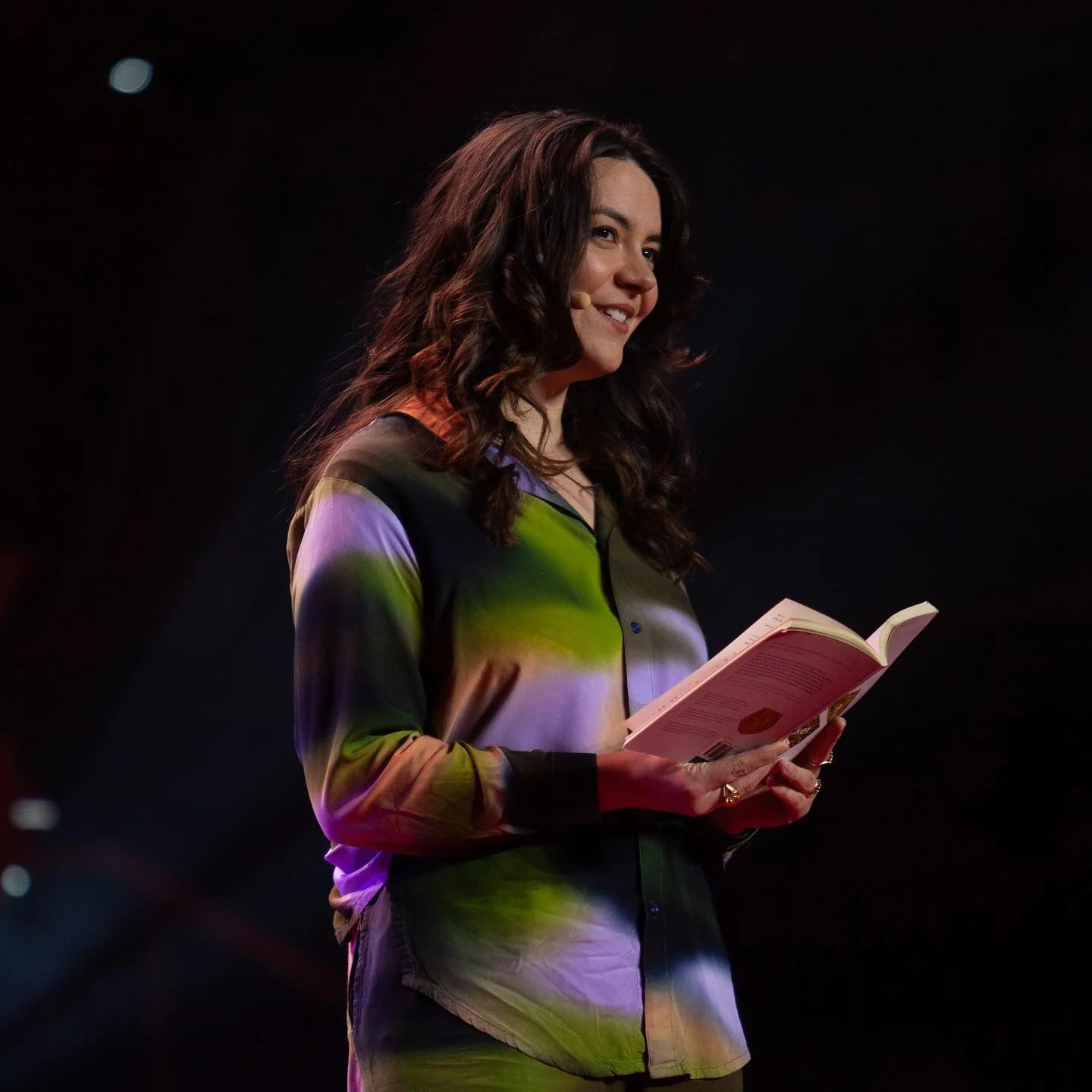 A woman with dark, curly hair holding an open book and smiling on stage, wearing a multicolored blouse.