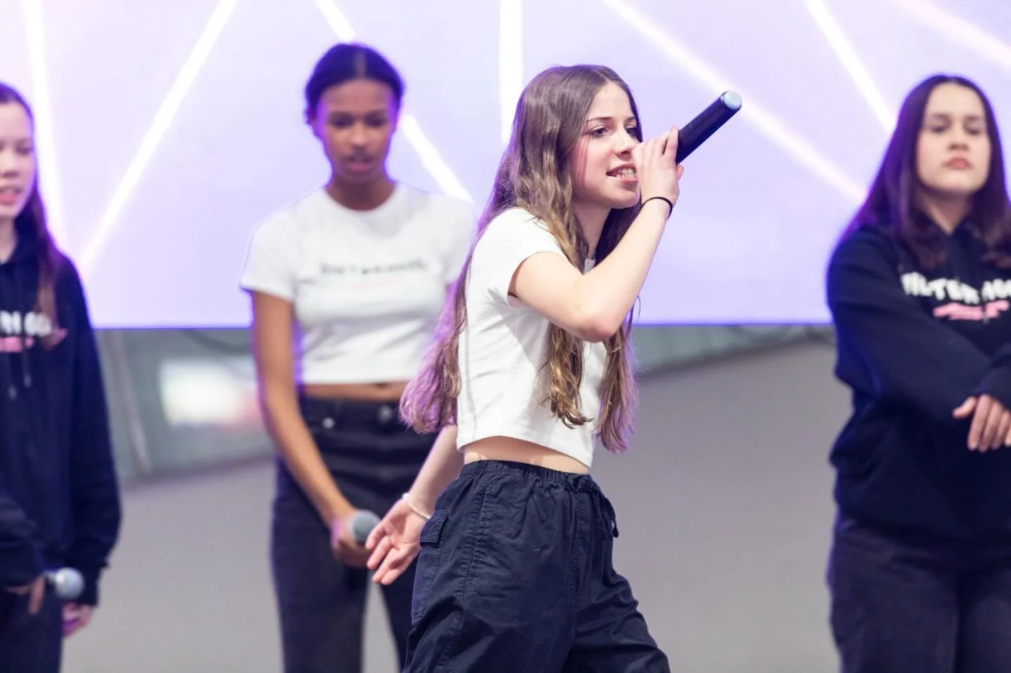 Young woman with long, wavy hair singing into a microphone on stage, surrounded by three other women in casual outfits.