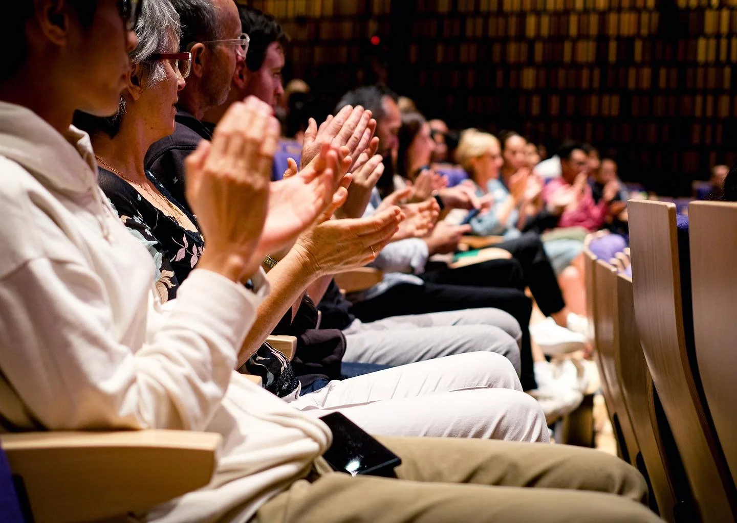 Audience members sitting in theater seats, clapping and watching a performance or presentation.