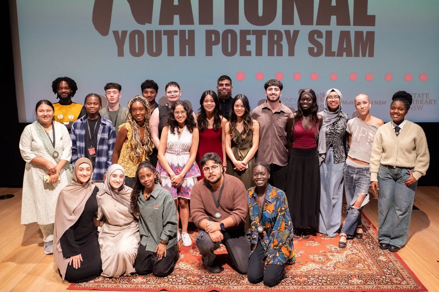 A diverse group of young people and adults posing on stage in front of a large screen that reads 'National Youth Poetry Slam.' They are smiling and standing together for a group photo. The stage has a wooden floor and a decorative rug in front of the group.
