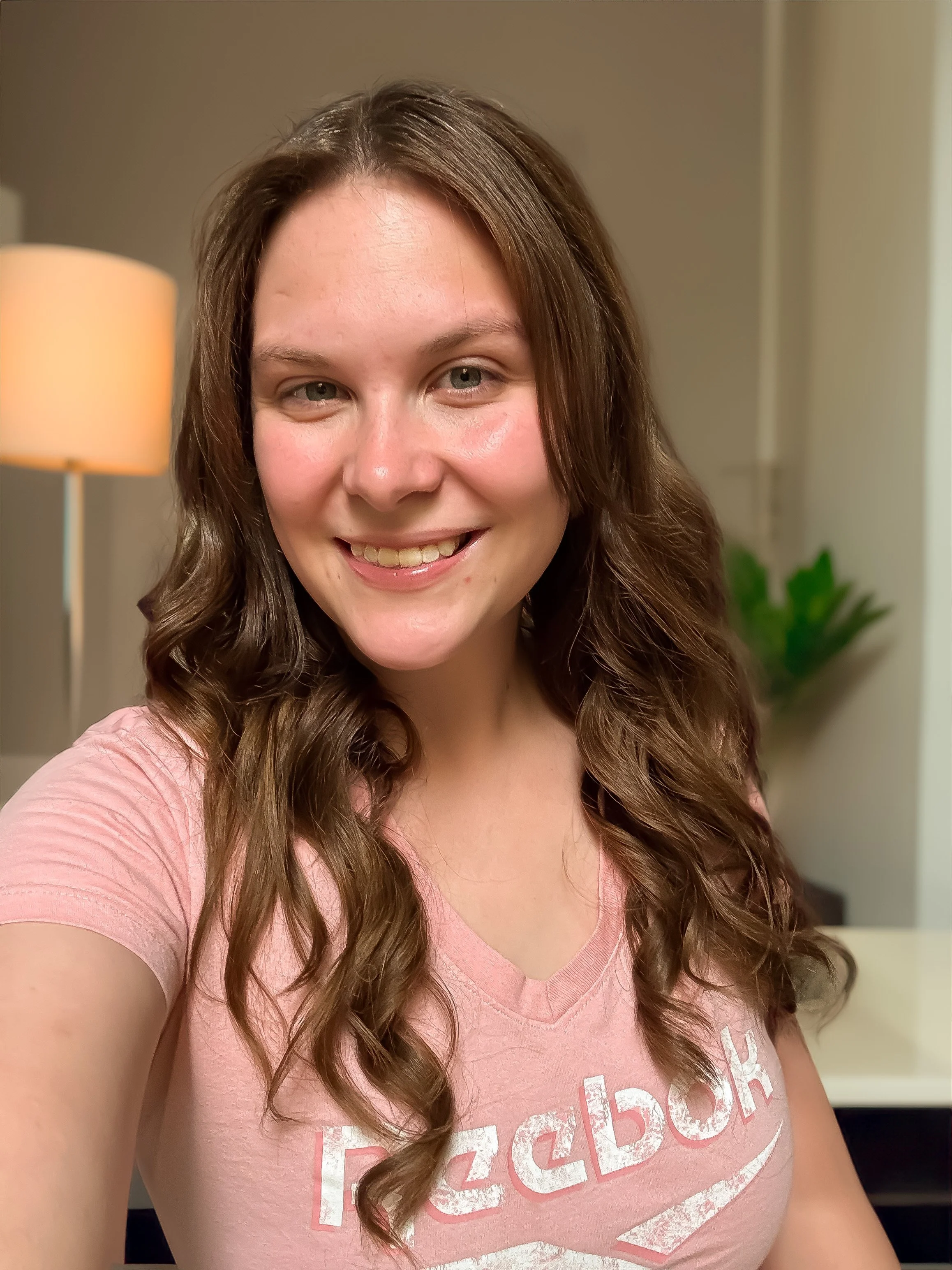 A young woman with long, wavy brown hair taking a selfie indoors, smiling, wearing a pink Reebok t-shirt, with a lamp and green plant visible in the background.