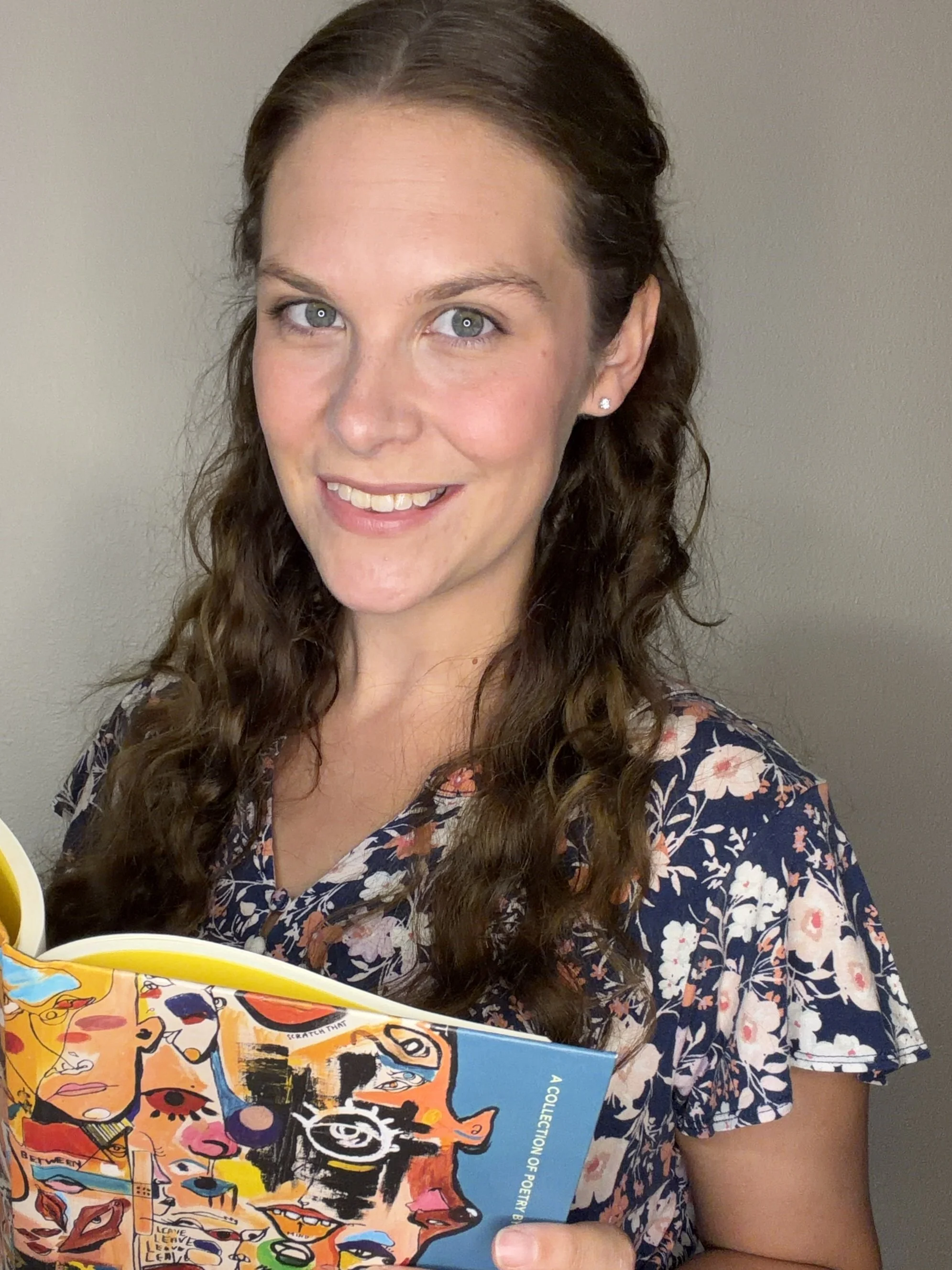 A young woman with long, curly brown hair, blue eyes, and fair skin, smiling and wearing a floral print shirt, holding a colorful book with abstract illustrations of faces in front of a plain wall.