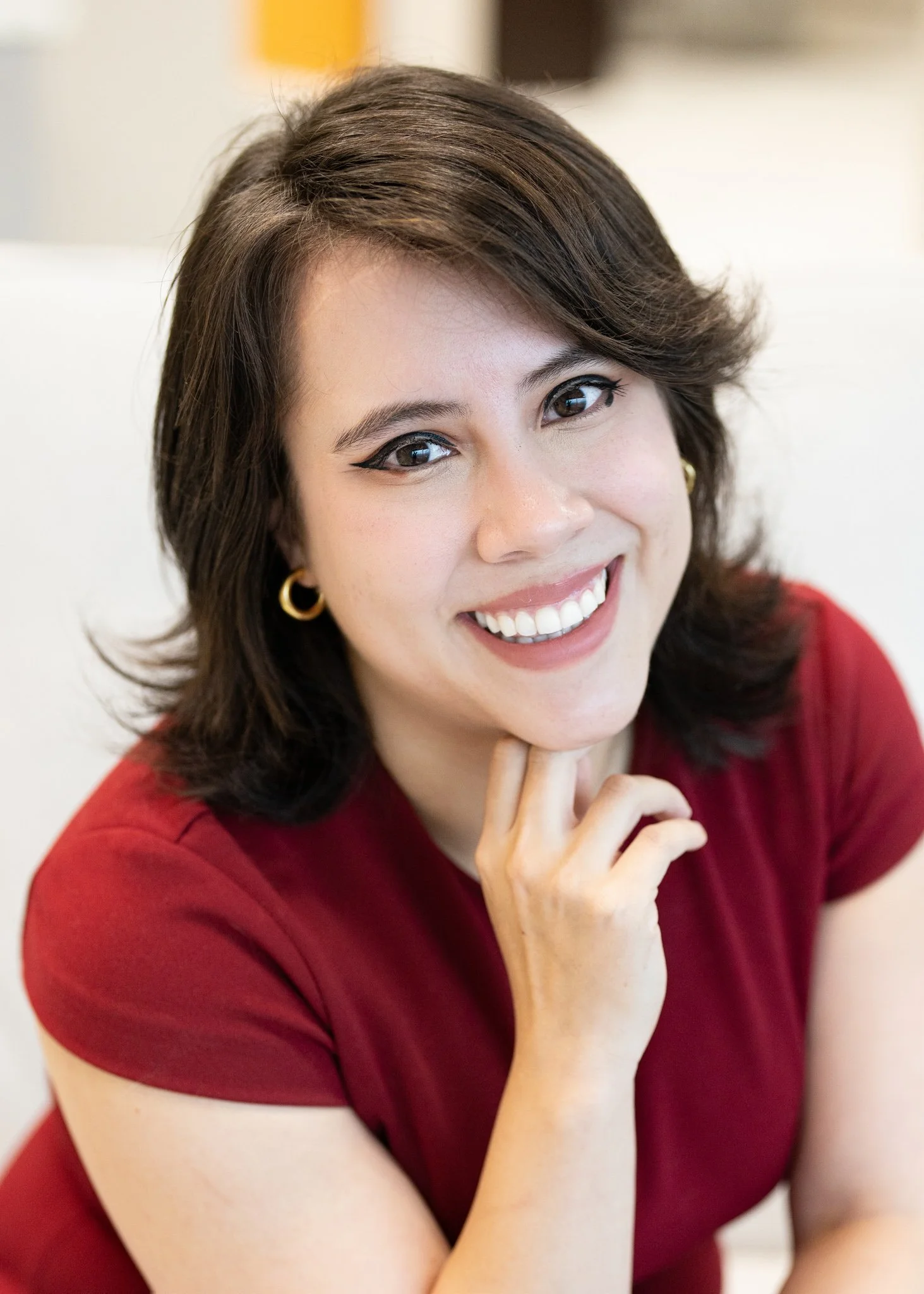 A woman with short dark hair, wearing a red top and gold earrings, smiling and looking at the camera with her hand resting under her chin.