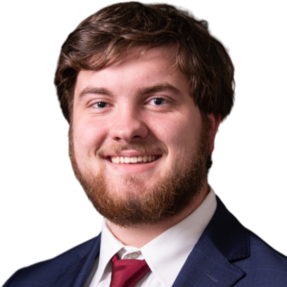 A young man with brown hair and a beard, smiling, dressed in a dark suit, white shirt, and red tie, against a plain white background.