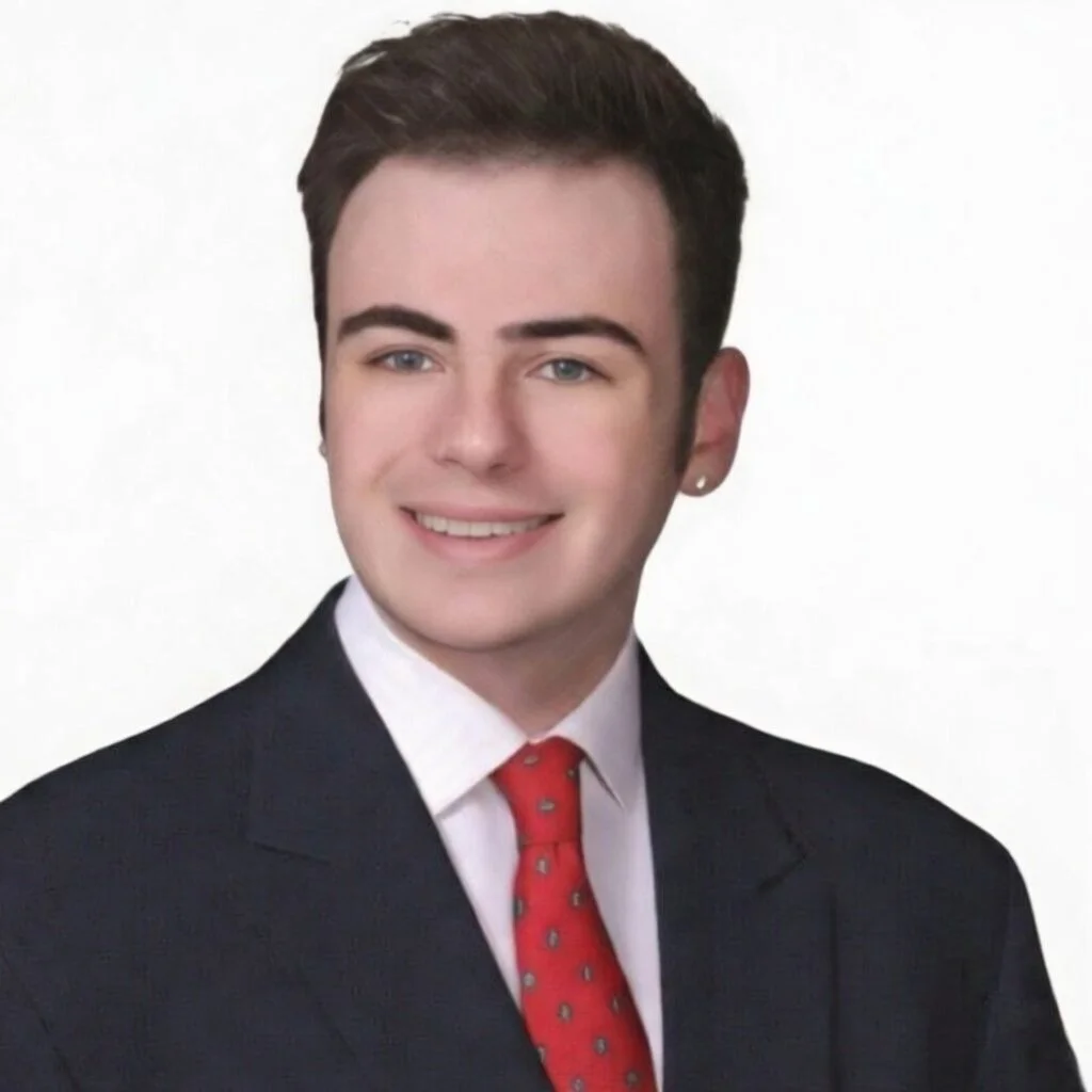 A young man with short dark hair, wearing a dark suit, white shirt, and red tie, smiling against a plain white background.