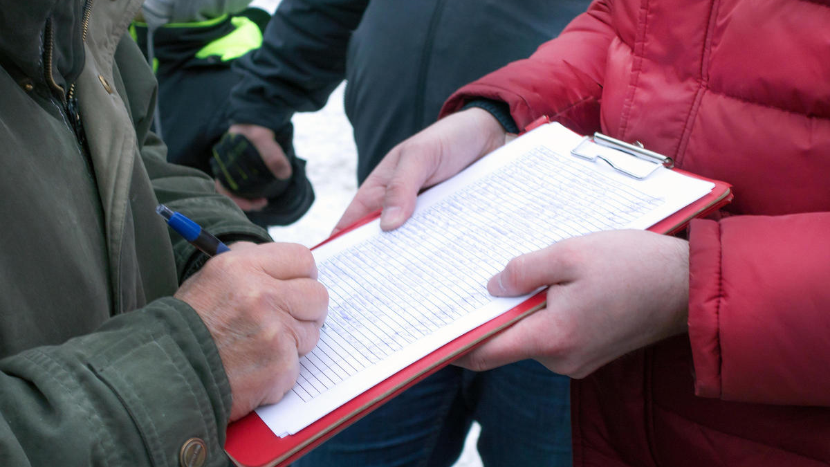 Two people exchanging a clipboard with handwritten notes outdoors, one signing with a pen and the other holding the clipboard.