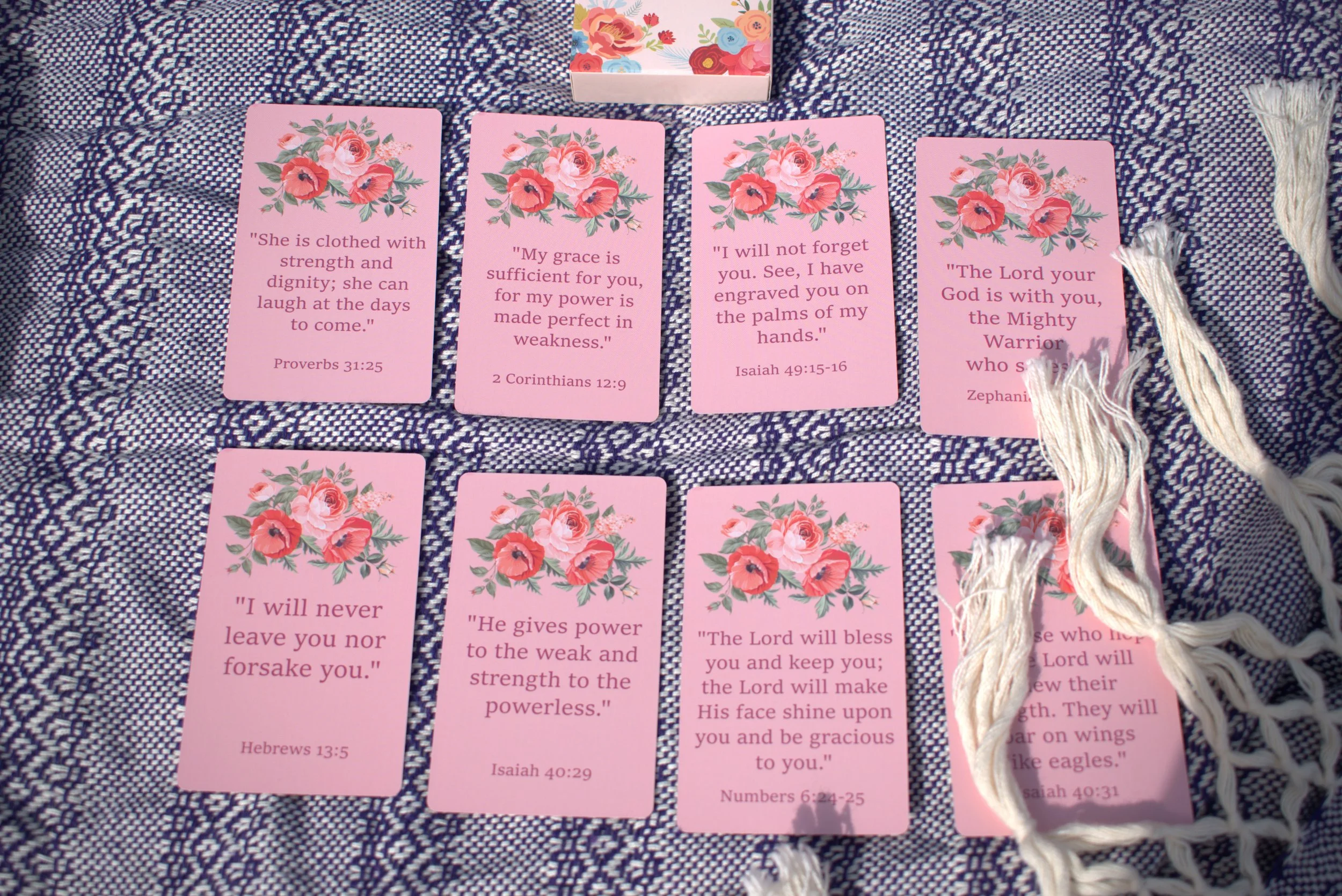 Pink prayer and motivational cards with floral designs and Bible quotes laid out on a blue patterned fabric. One card is partially visible at the top, and a white tassel or fringe hangs from the right side.