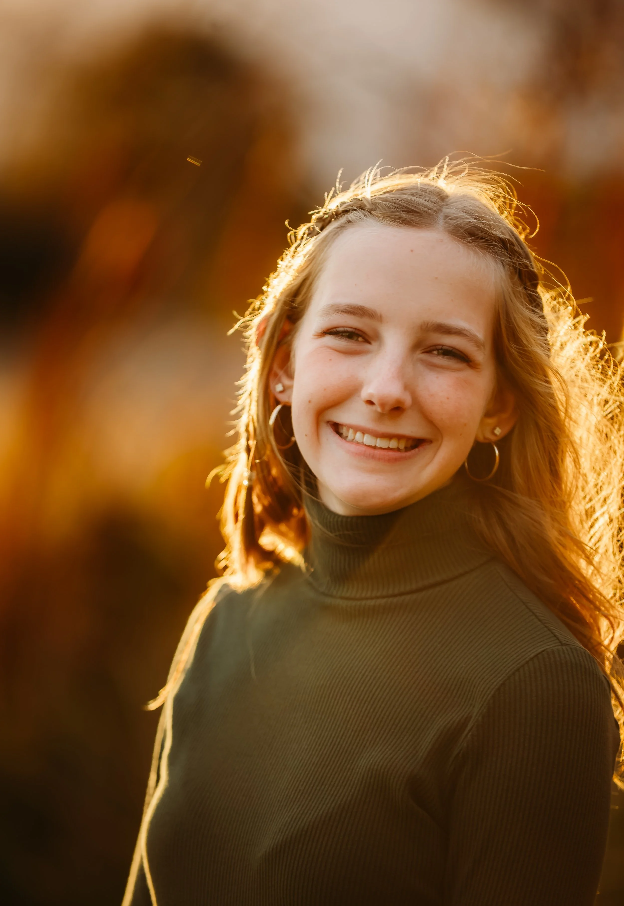 Close-up of a young woman smiling outdoors during golden hour sunlight, with blurred autumn foliage in the background.
