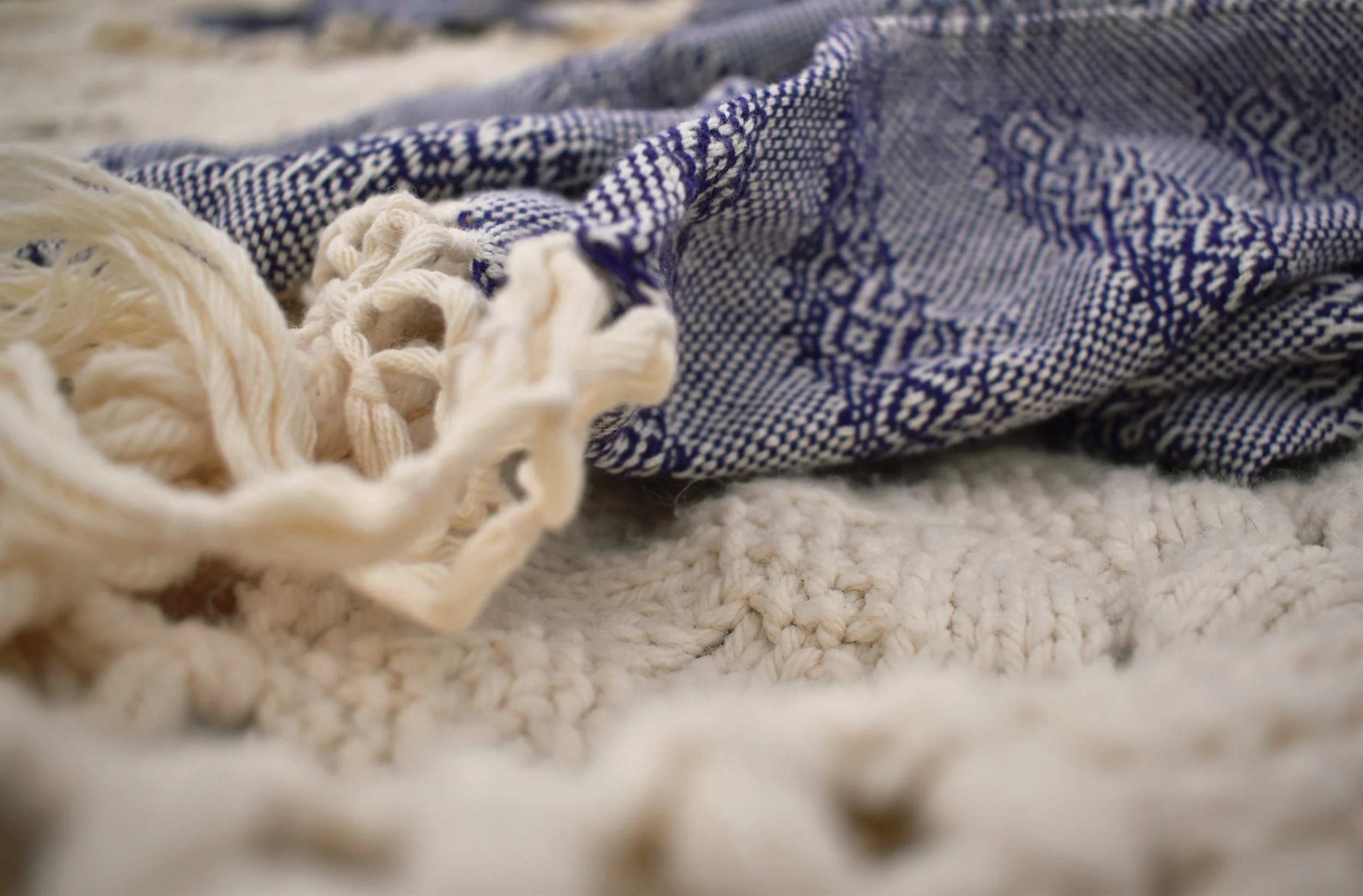 A close-up of a patterned blue and white textile with fringe, resting on a cream-colored textured surface.