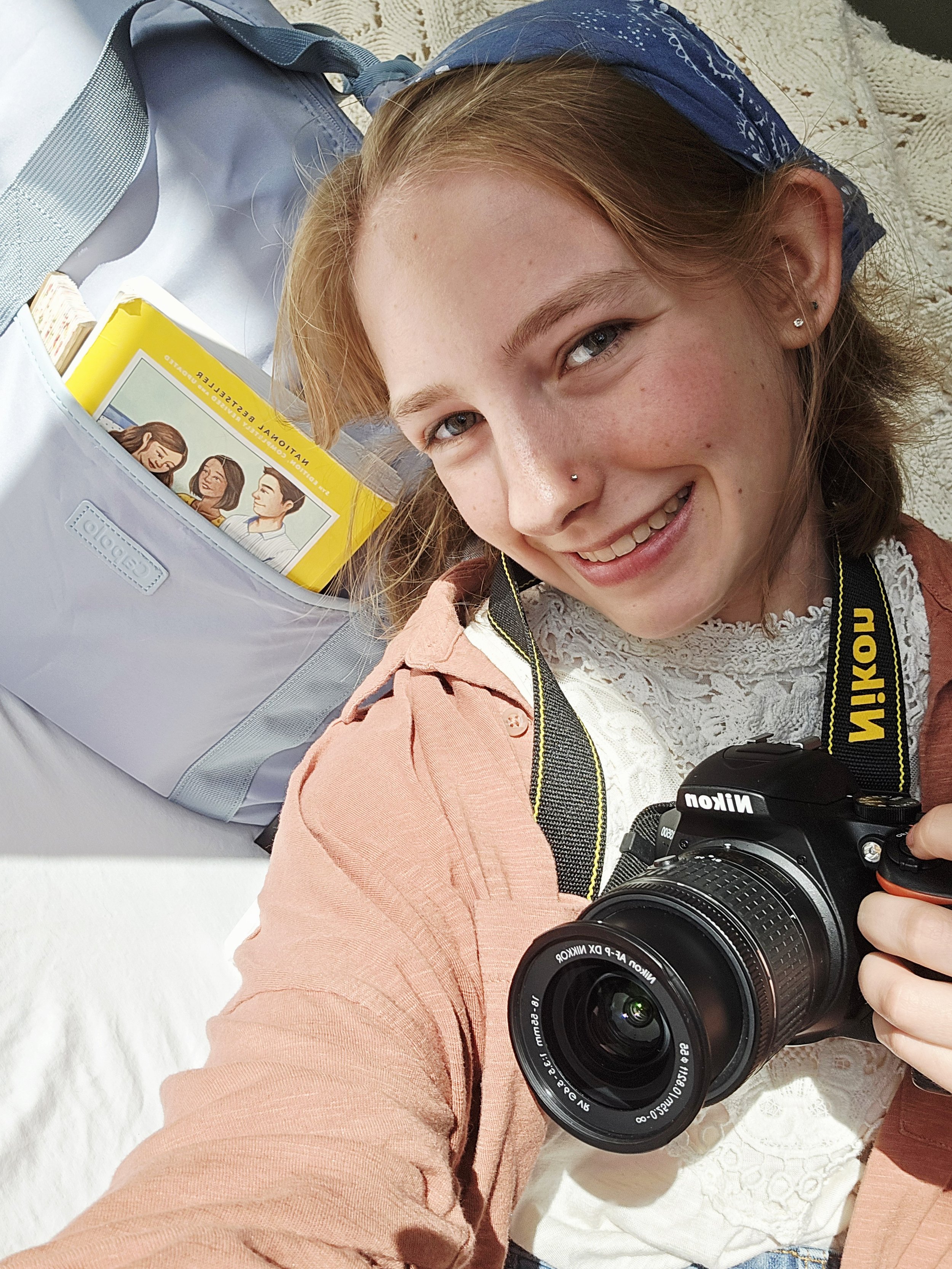 A young woman with red hair, light skin, and facial piercings taking a selfie with a Nikon DSLR camera. She is smiling and wearing a blue bandana, a white lace top, and a peach-colored jacket. There is a white bag with a book inside and a blue strap on her shoulder.