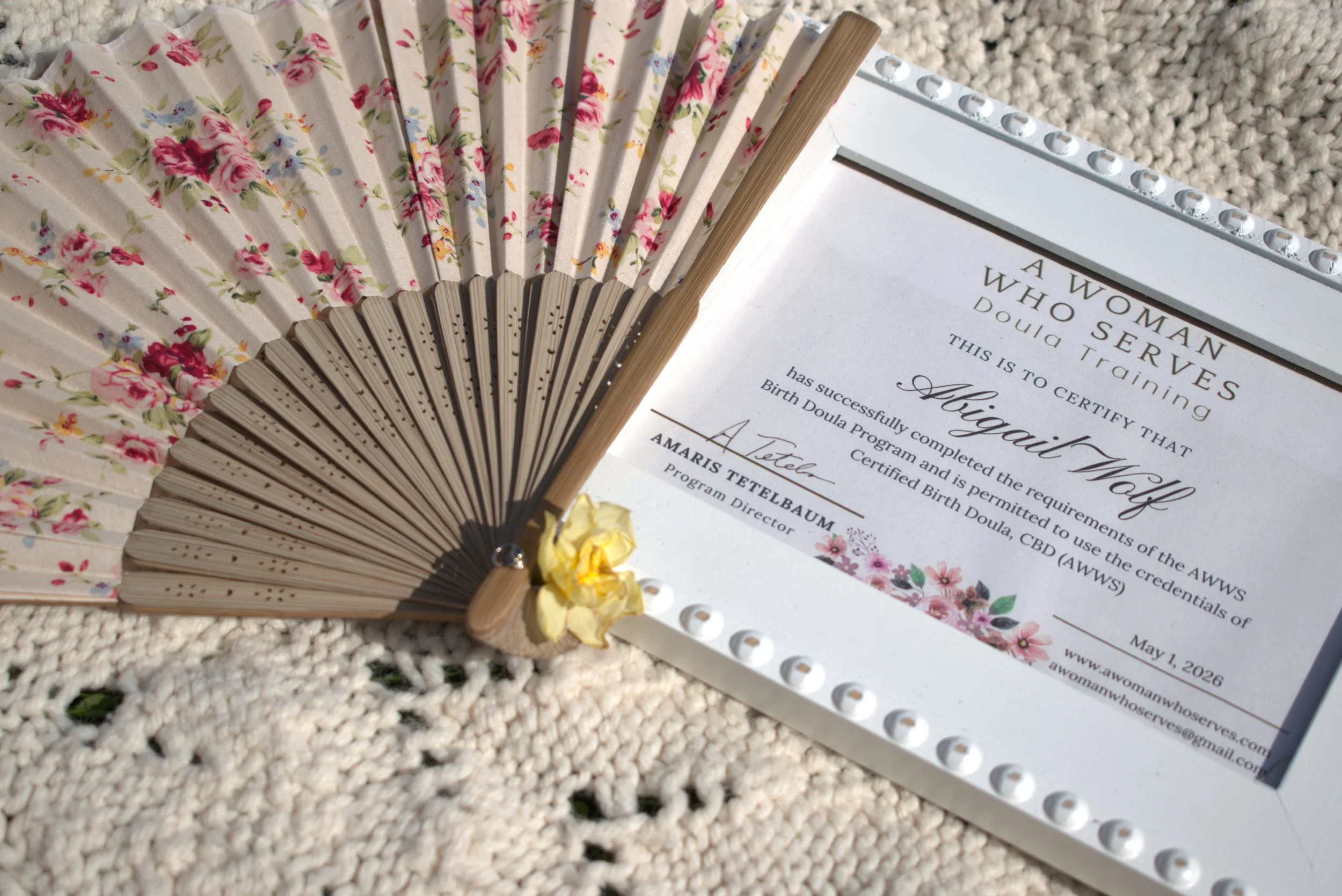 A floral patterned hand fan resting near a certificate of completion for a program called 'A Woman Who Serves', with a textured beige fabric background.