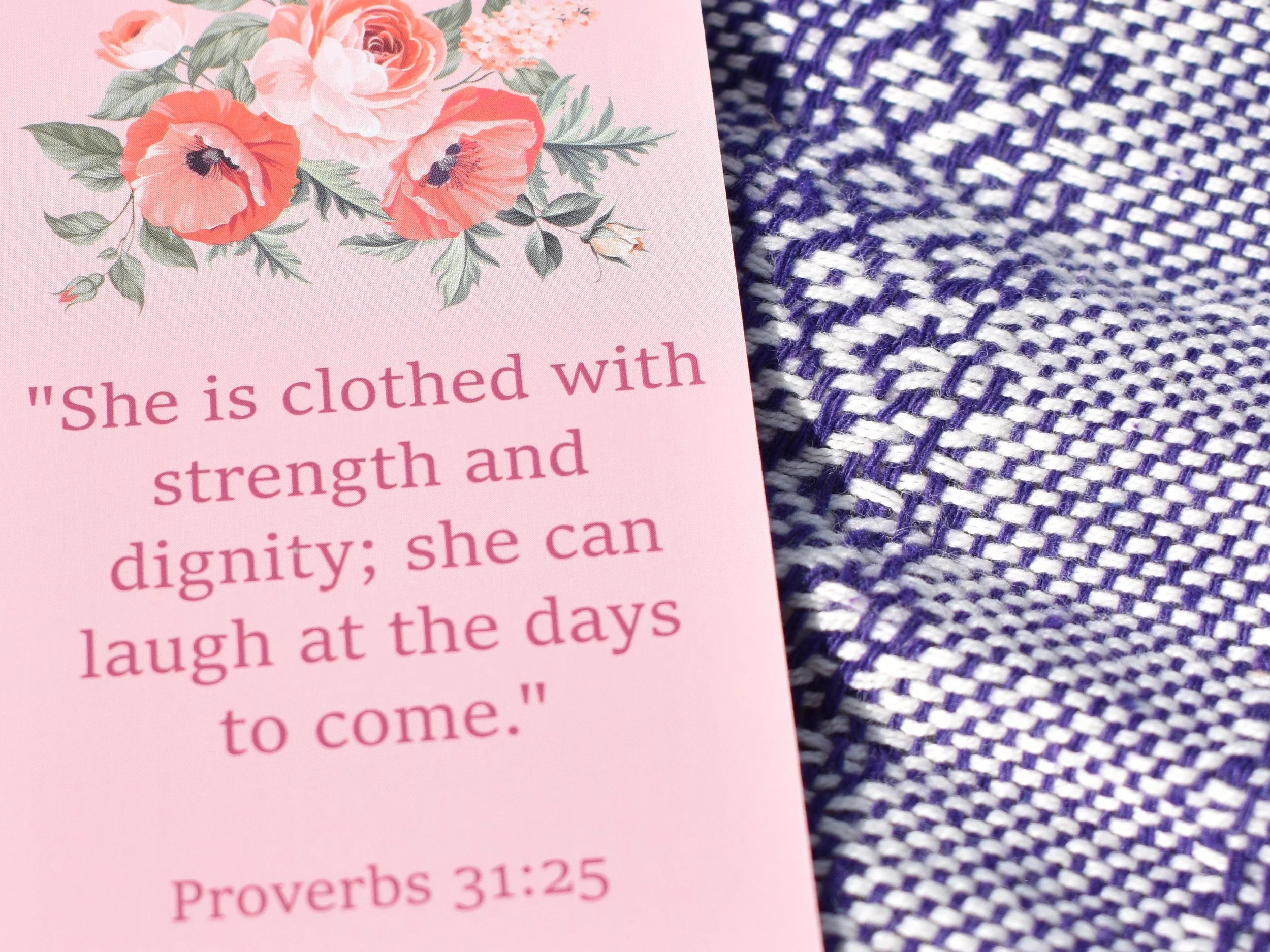 Close-up of a pink card with a floral design and partial quote about strength and dignity, placed on a woven blue and white fabric background.