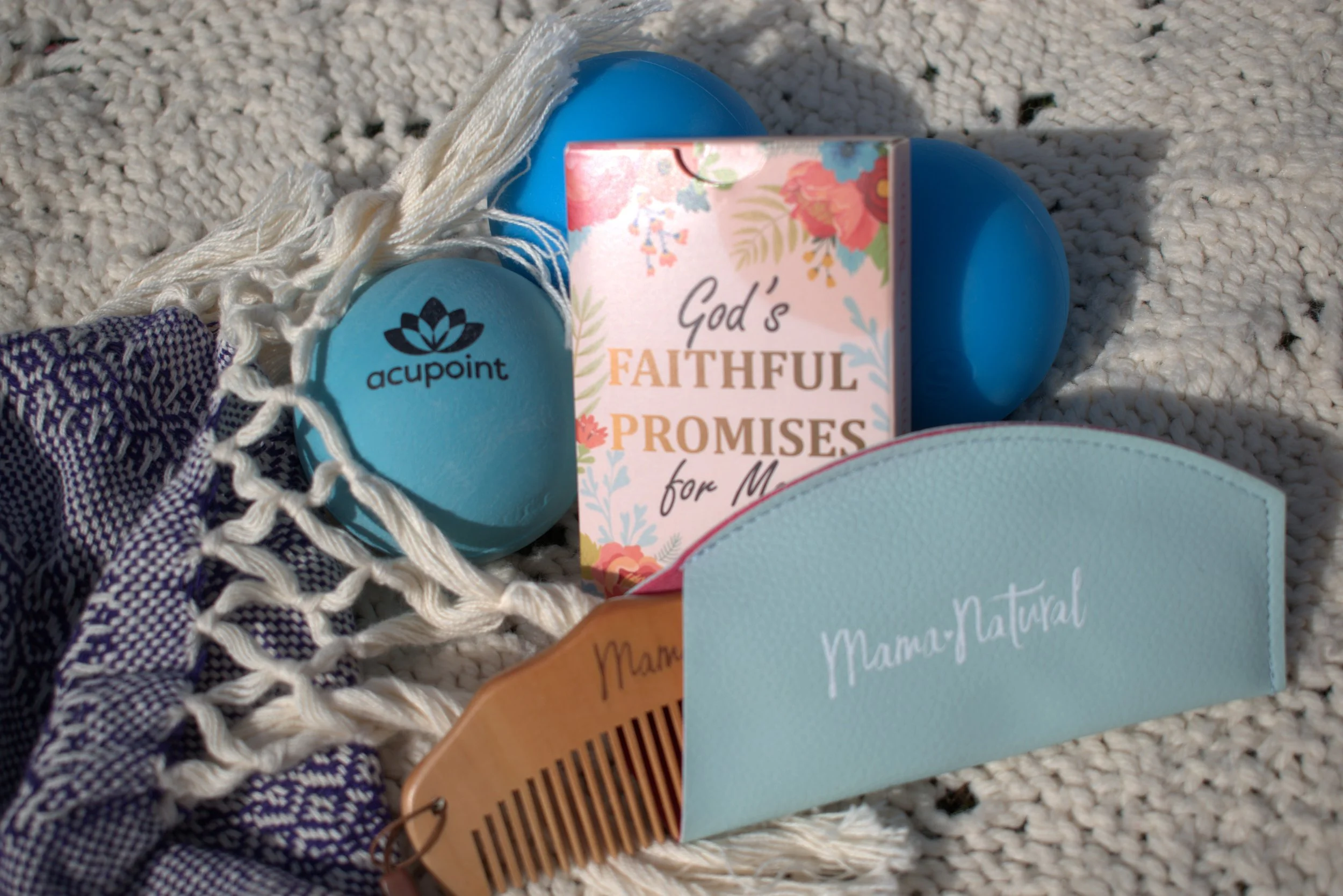 Collection of various personal items including a blue balloon, a pink floral card with the words "God's Faithful Promises for M...," a light blue card with white text, a woven hair comb, a piece of purple and white fabric, and a cream-colored decorative net on a textured white surface.