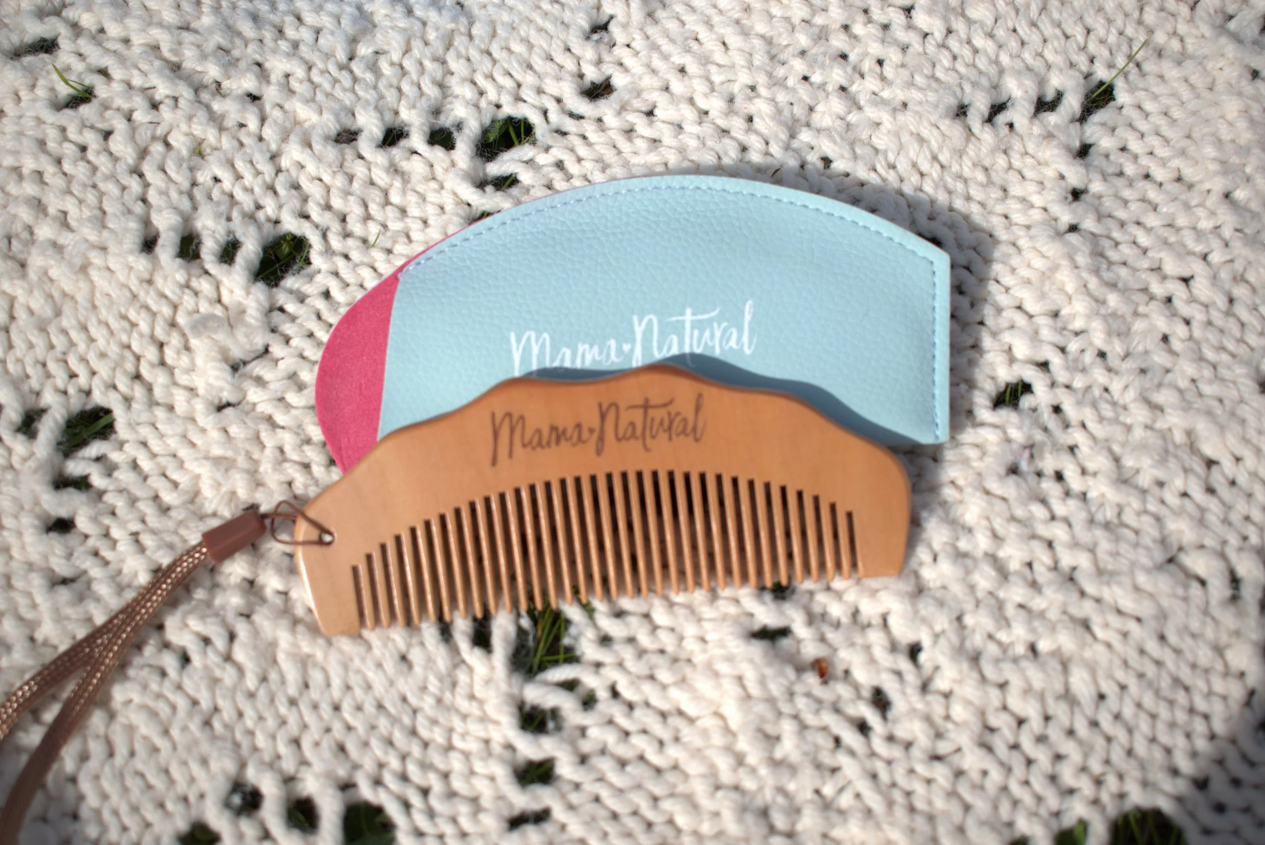 A wooden hairbrush with a curved handle and wide spaced teeth on a woven white outdoor rug, with a blue, pink, and brown paper tags attached to it.