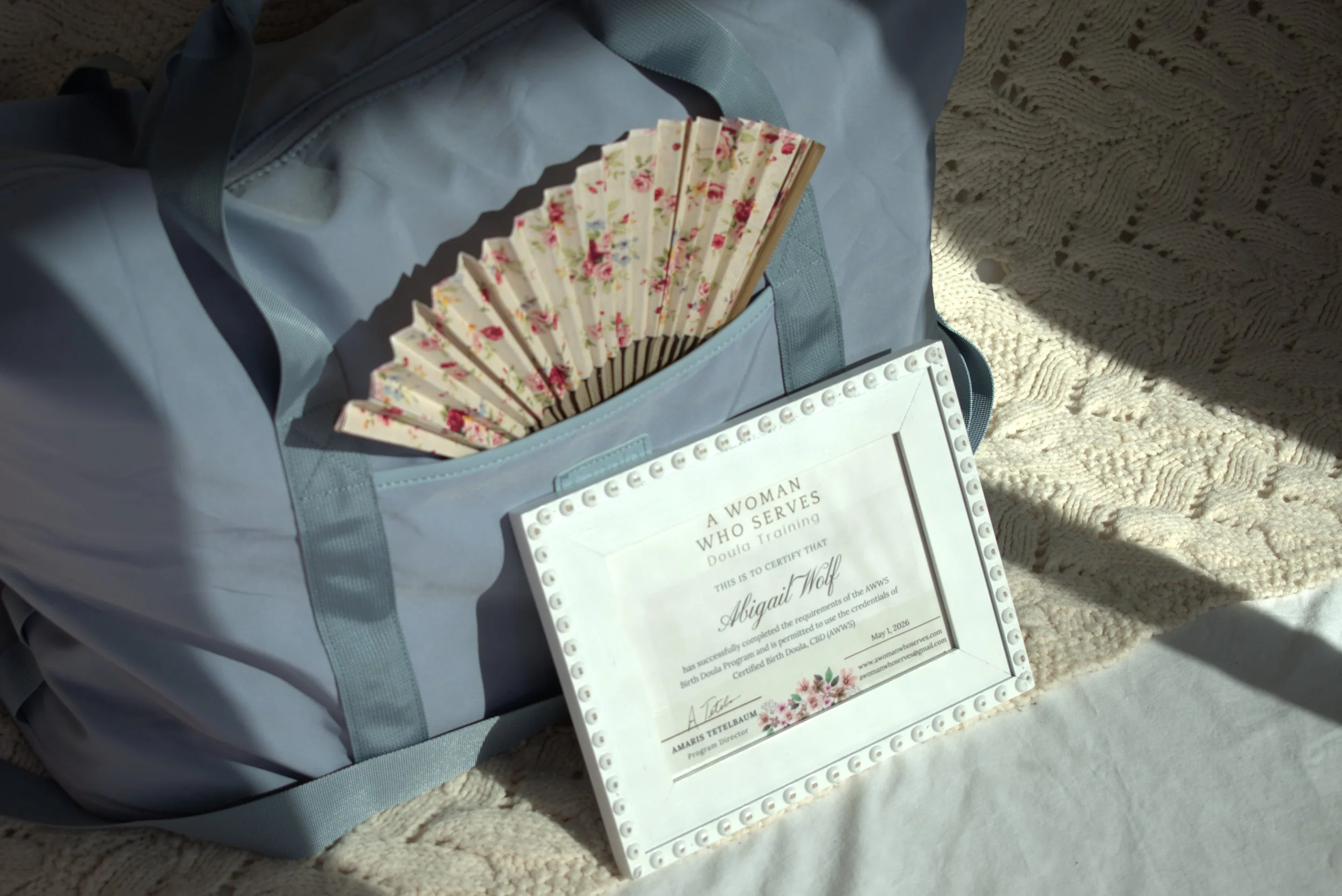 A light blue bag with a fan, a certificate, and a whiteboard inside, sitting on a crocheted blanket with sunlight coming through.