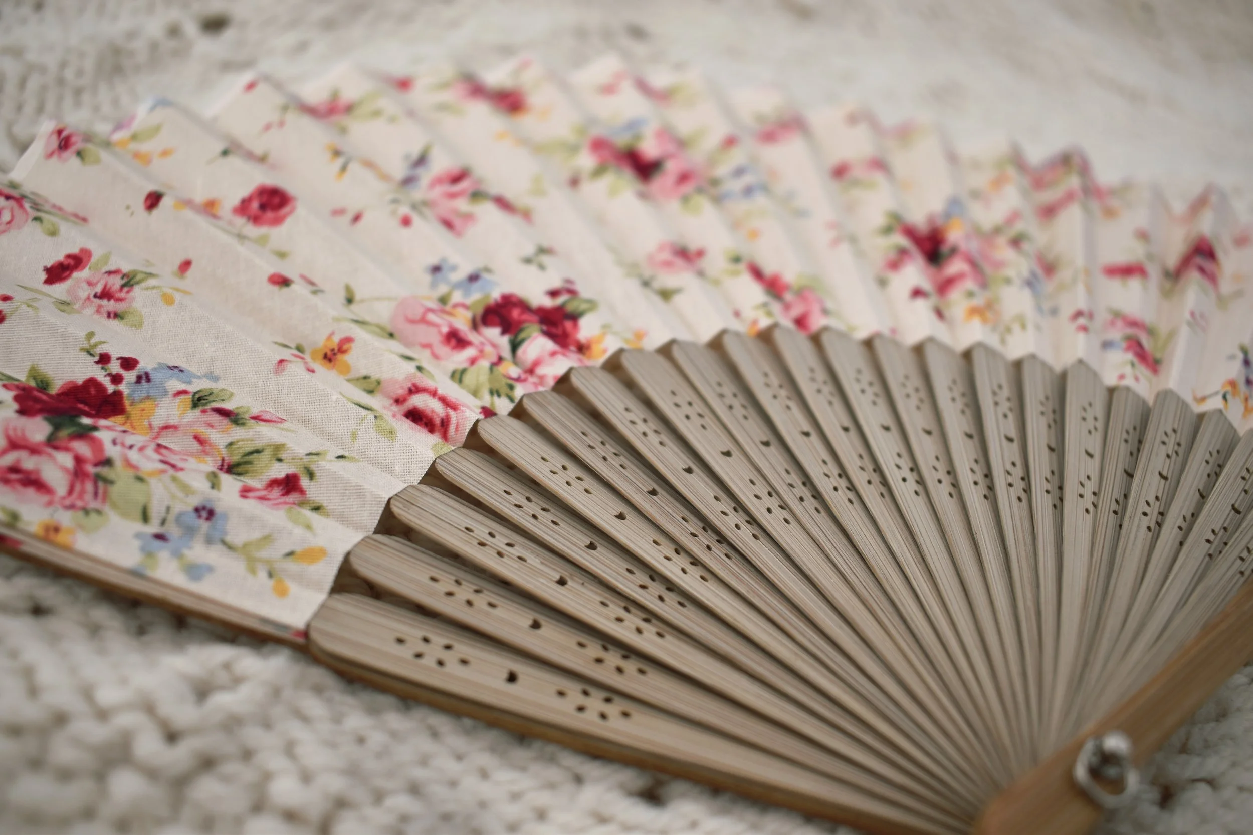A decorative folding paper fan with a floral pattern of pink roses and greenery, lying open on a textured light-colored surface.