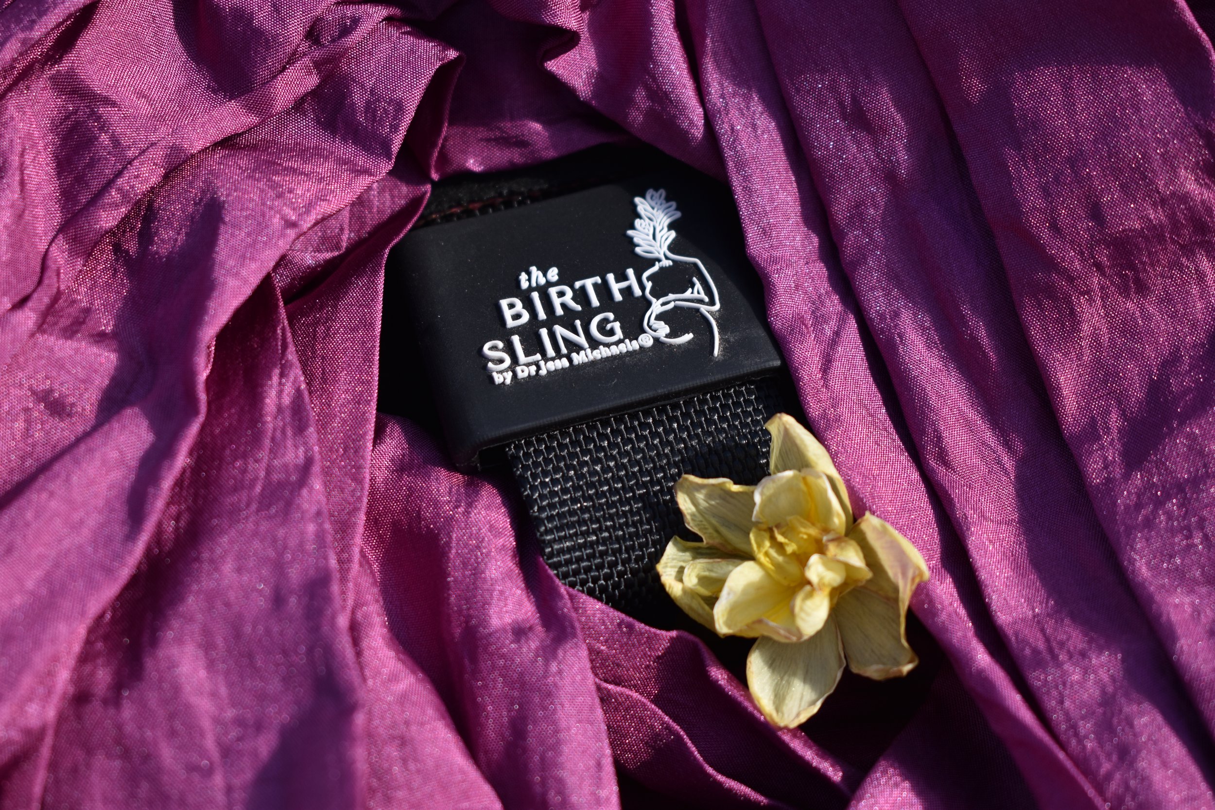 A black fake belt with a white label reading 'the BIRTH SLING by Dr. Jean Meleshe' surrounded by purple shiny fabric and yellow dried flowers.