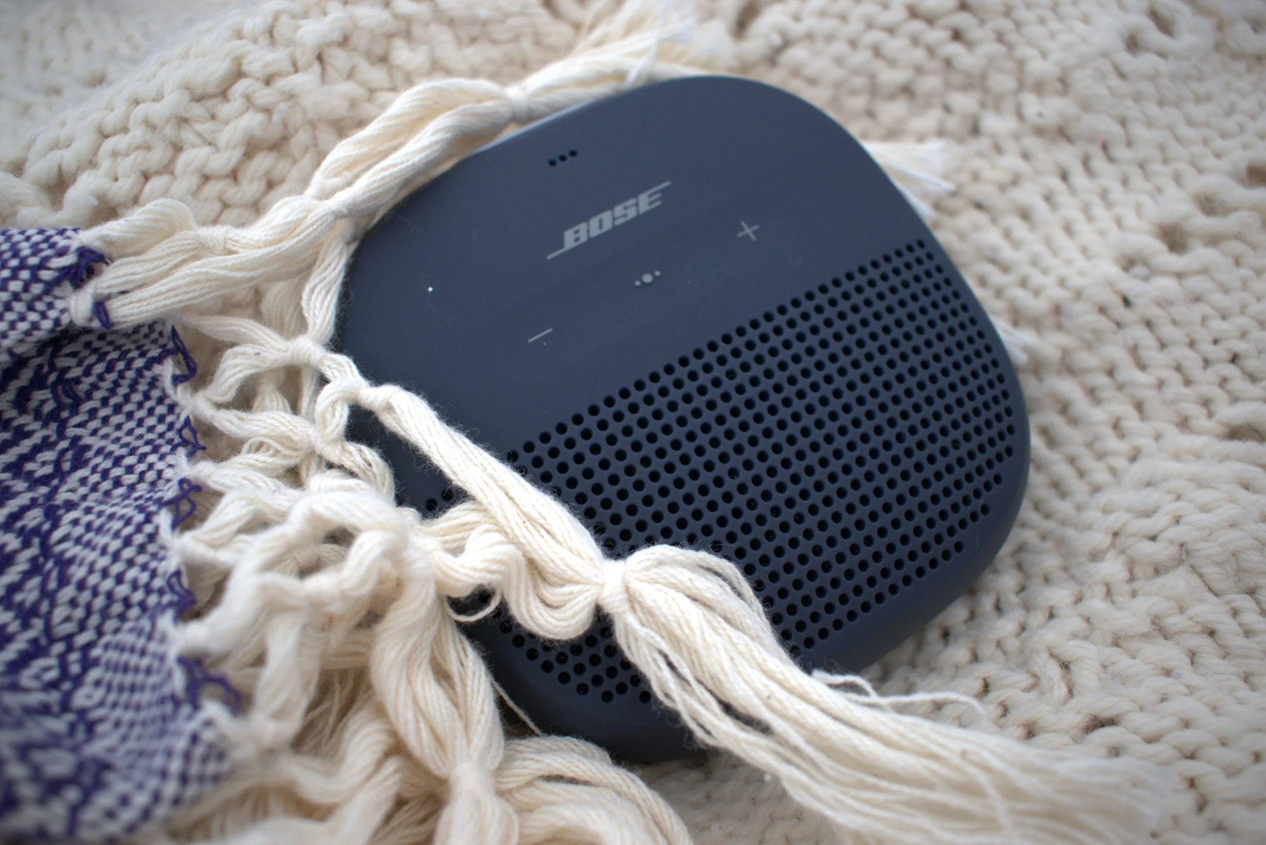 Close-up of a black Bose portable speaker placed on cream-colored, chunky knit blanket with a piece of woven fabric and twisted cord on top.