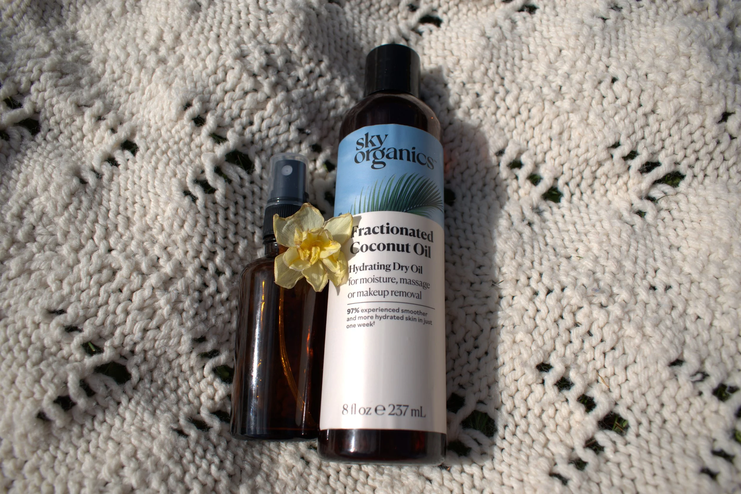 A bottle of Sky Organics Fractionated Coconut Oil with a yellow flower and a smaller spray bottle placed on a white textured blanket.