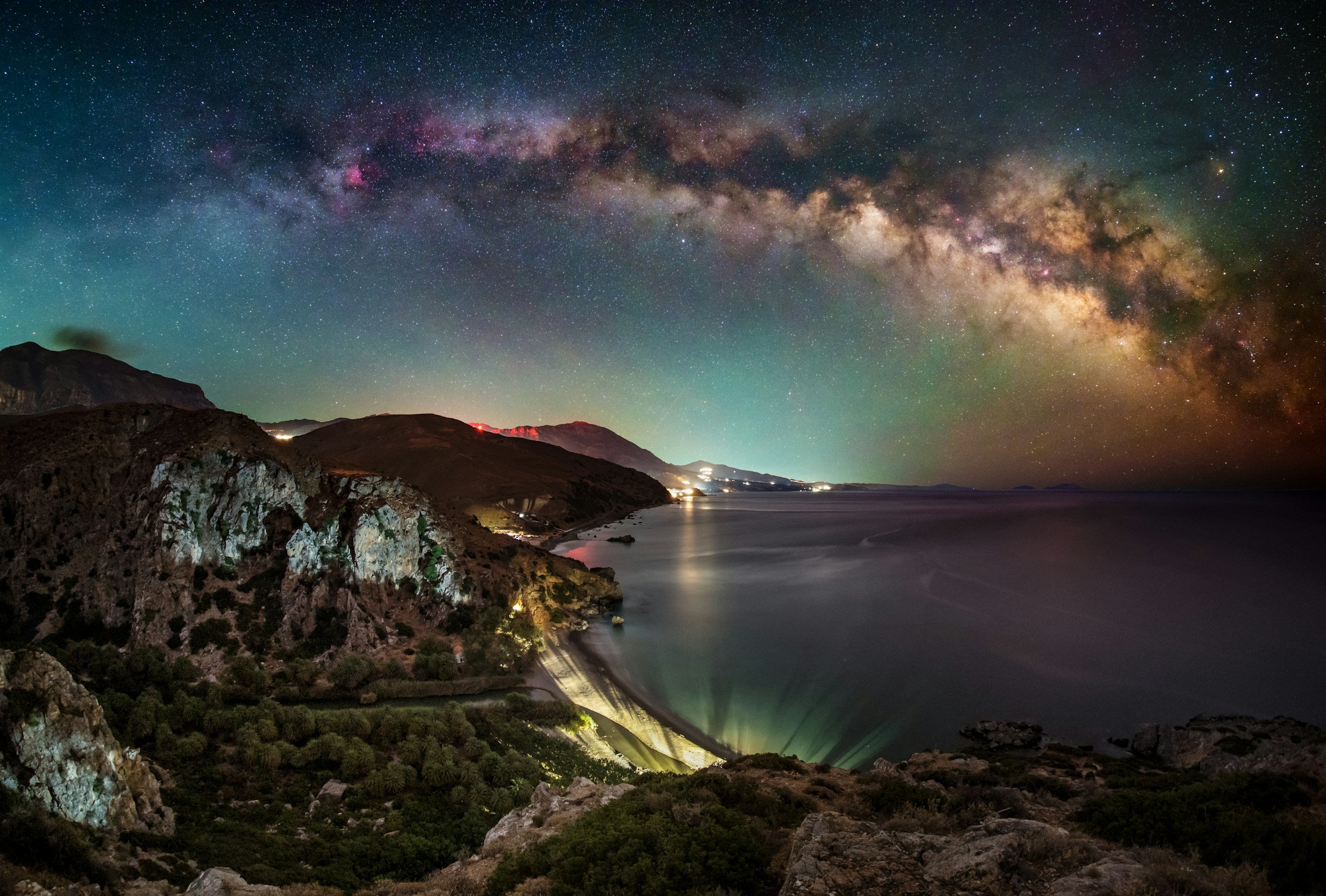 Nighttime landscape showing rocky coastline and a calm body of water under a starry sky with the Milky Way galaxy visible.