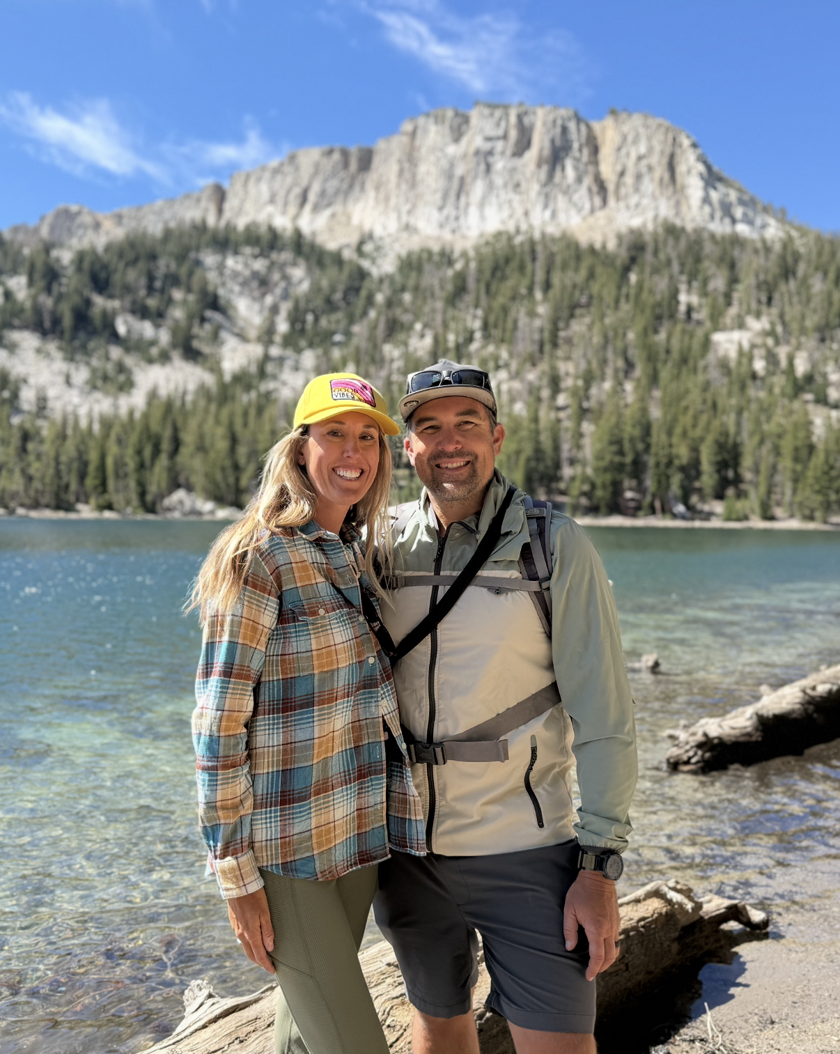 A smiling couple standing on a rocky shore in front of a lake with a mountain and a forested area in the background, enjoying a sunny day outdoors.