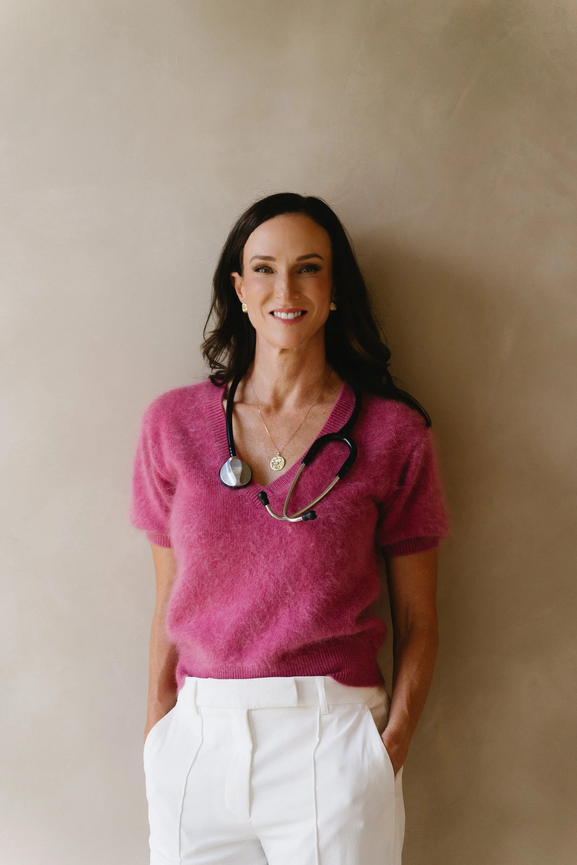 Woman in pink top and white pants smiling. She has a stethoscope around her neck.