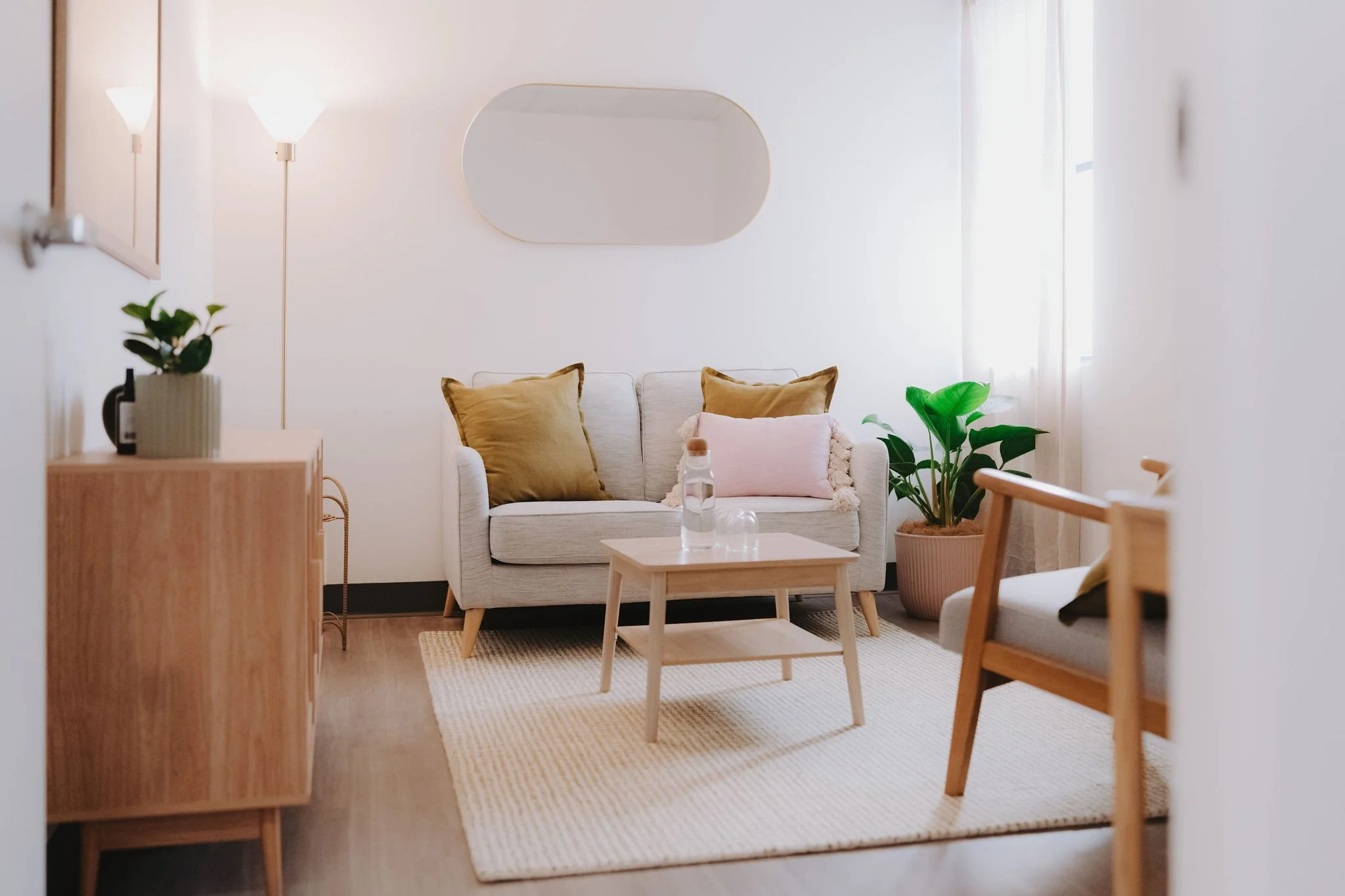 Cozy living room with a white sofa, brown and pink pillows, a small wooden coffee table with water bottles, a large potted plant, a woven rug, and ambient lighting from two wall lamps and a window with curtains.