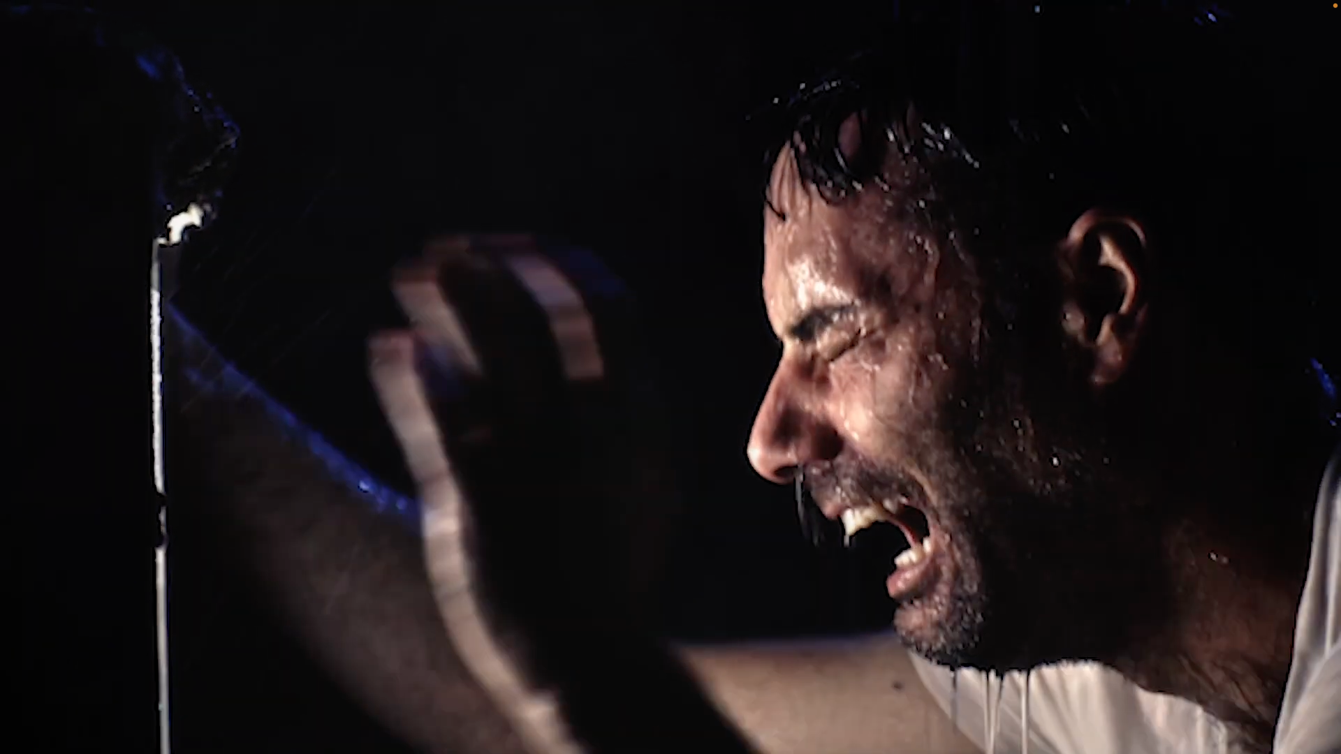 A man with wet hair and a beard screams with his eyes closed, face contorted in pain or anger, in a dark setting.