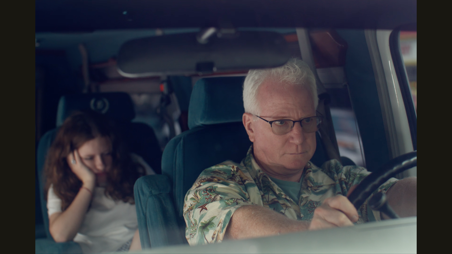 An older man with white hair and glasses driving a vehicle, with a young girl with curly hair sitting behind him, holding her ear.