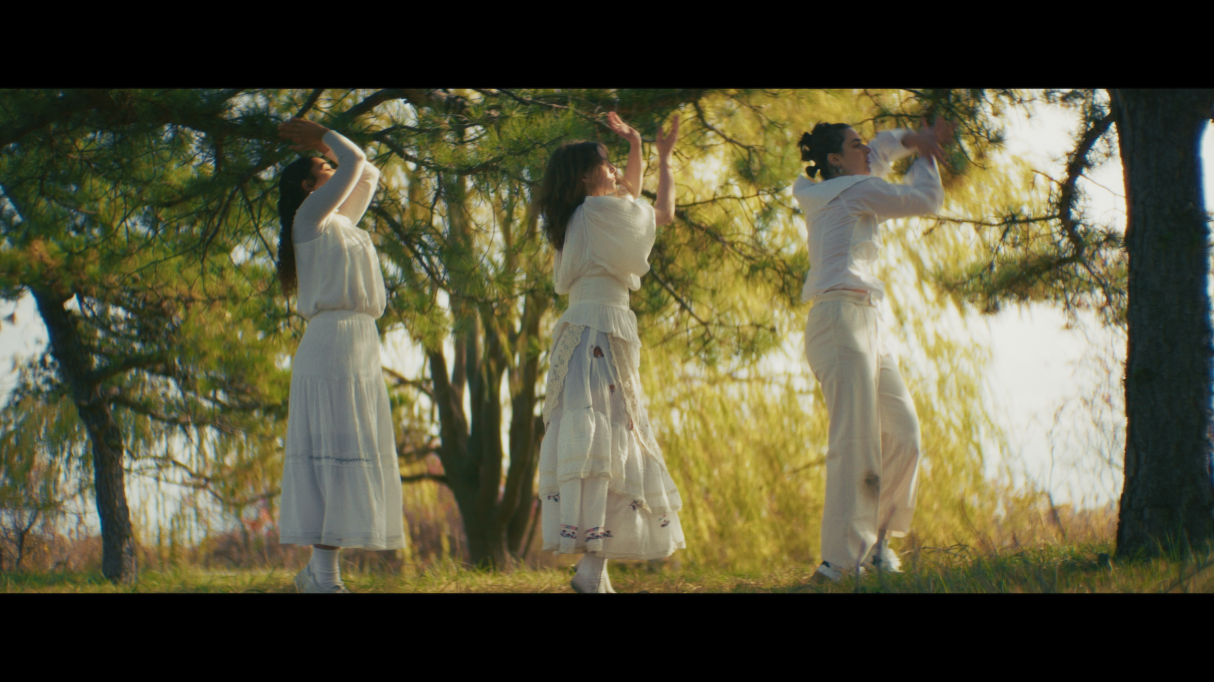 Three women in white dresses dancing outdoors under trees on a sunny day.
