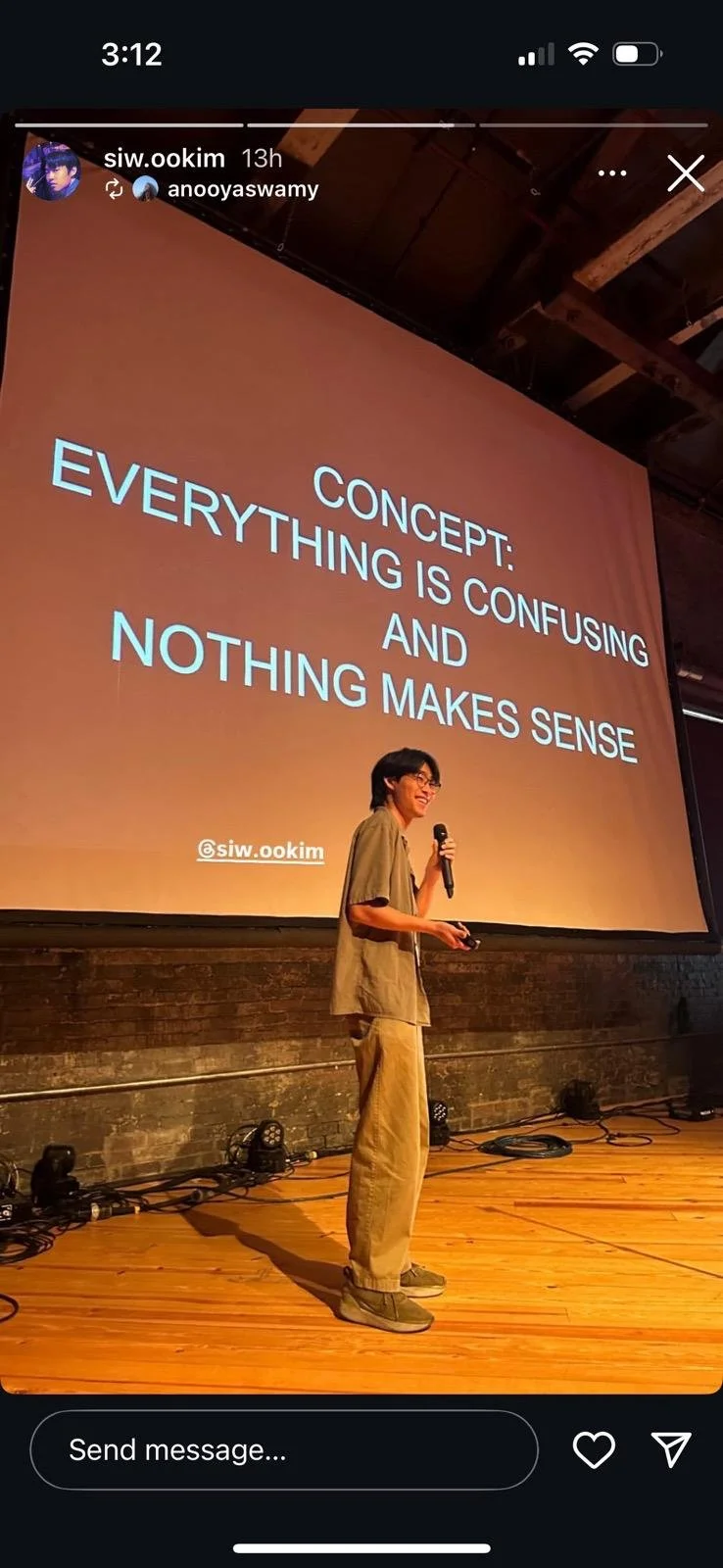 A person holding a microphone and a remote control stands on a wooden stage in front of a large screen displaying a presentation slide that reads: 'Concept: Everything is confusing and nothing makes sense'.