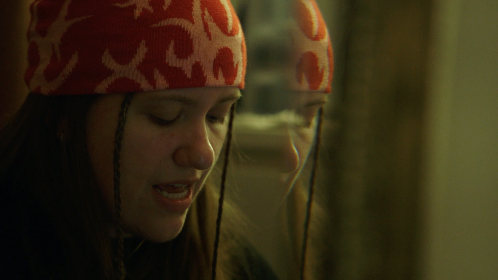 A young woman wearing an orange and white patterned headscarf, looking downwards, with her reflection visible in a mirror.