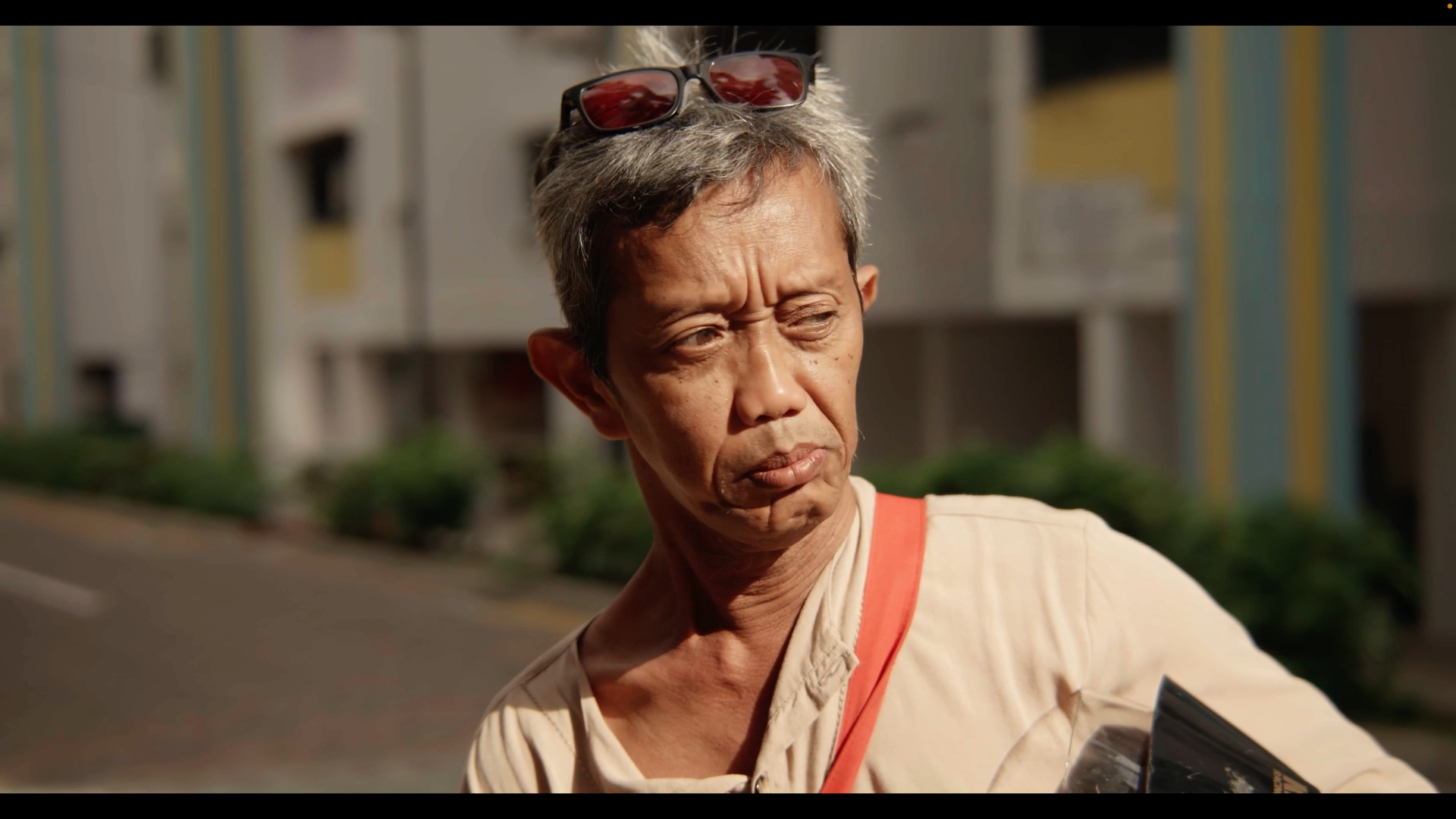 An older man with short gray hair, wearing sunglasses on his head, looking to the side with a concerned or confused expression, holding a black object in his hand outdoors with a blurred background of buildings and greenery.