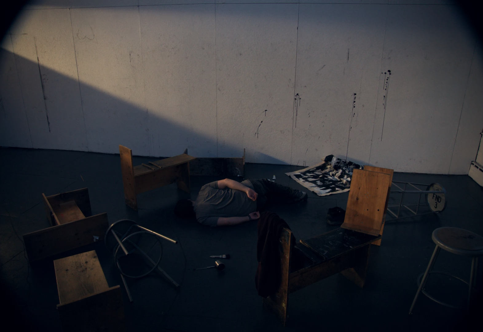 A person lying on the floor in a dark room with scattered furniture, including chairs and a small table, and a painted backdrop on the wall.