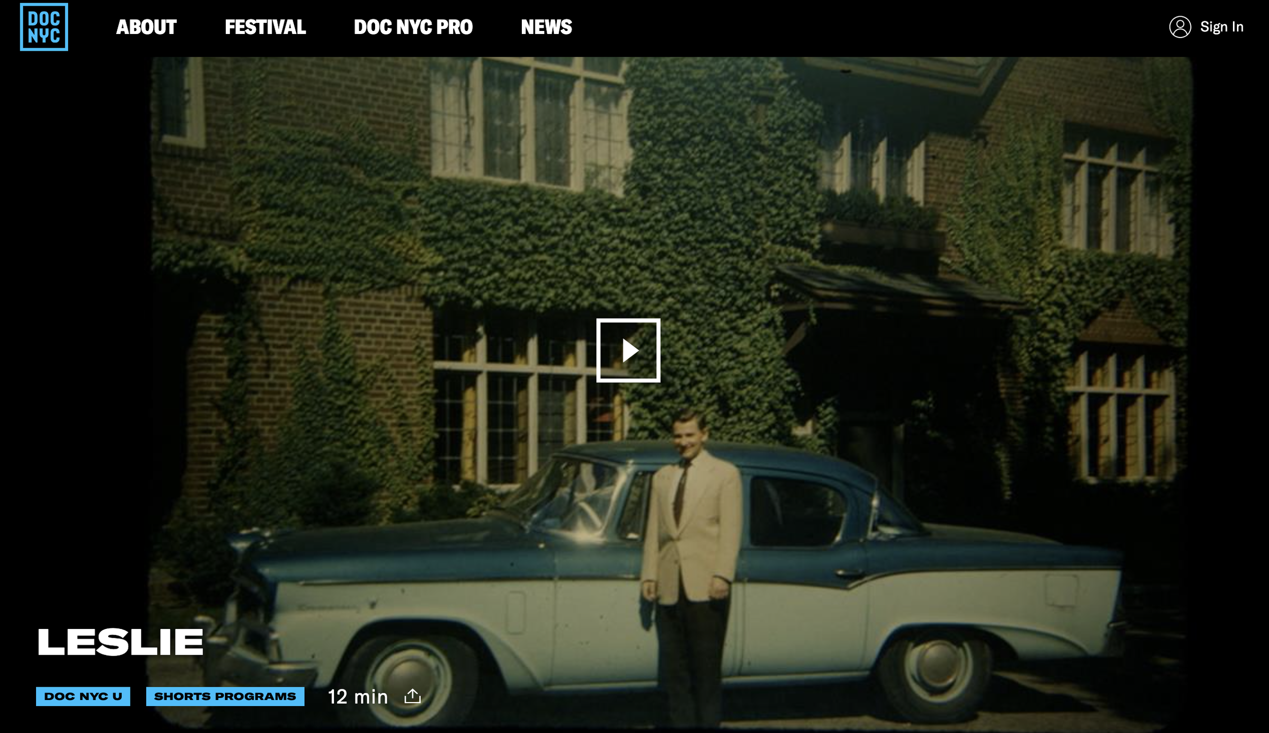 A man in a beige suit standing next to a vintage blue and white car in front of a brick house with ivy-covered walls and large windows.