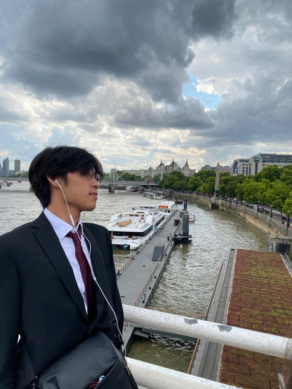 A young man in a black suit and white shirt stands near a river, listening to music with earphones, with boats on the river and a cityscape with cloudy skies in the background.