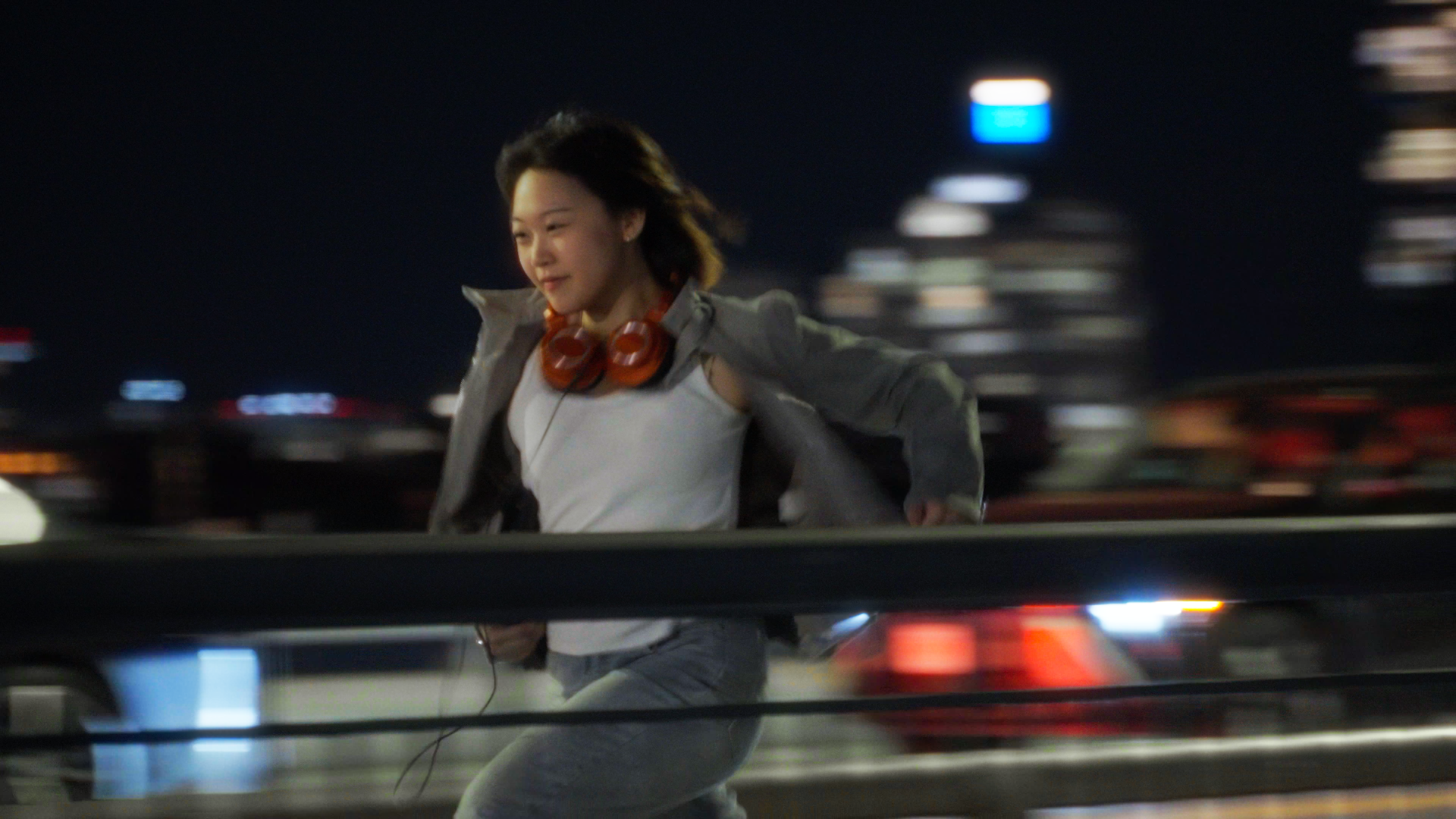A young woman running at night in an urban environment with city lights in the background, wearing headphones around her neck and a gray jacket.