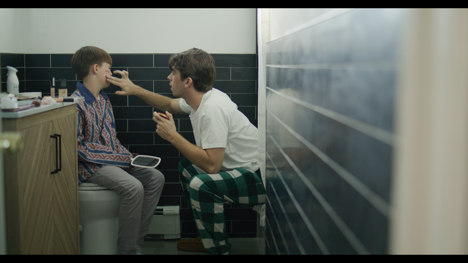 A man and a boy sitting on a toilet in a bathroom, with the man touching the boy's nose and holding a phone. The bathroom has black tiled walls.