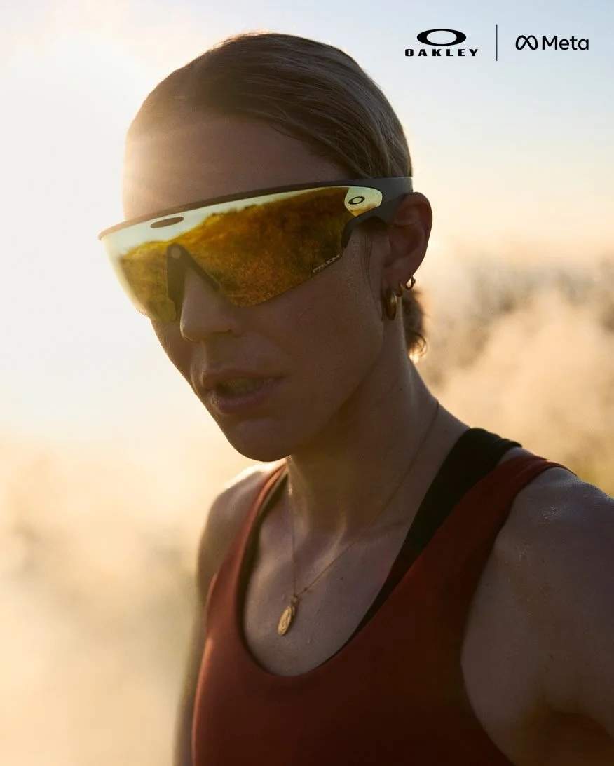 A woman wearing reflective sports sunglasses, a red sports top with black straps, and gold jewelry, outdoors during sunset.