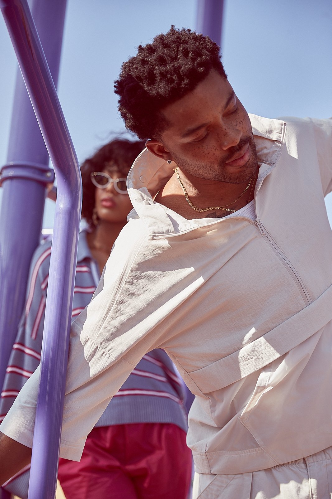 A man with short, curly hair and a woman with curly hair and sunglasses standing outdoors under a clear blue sky, near a purple playground structure.