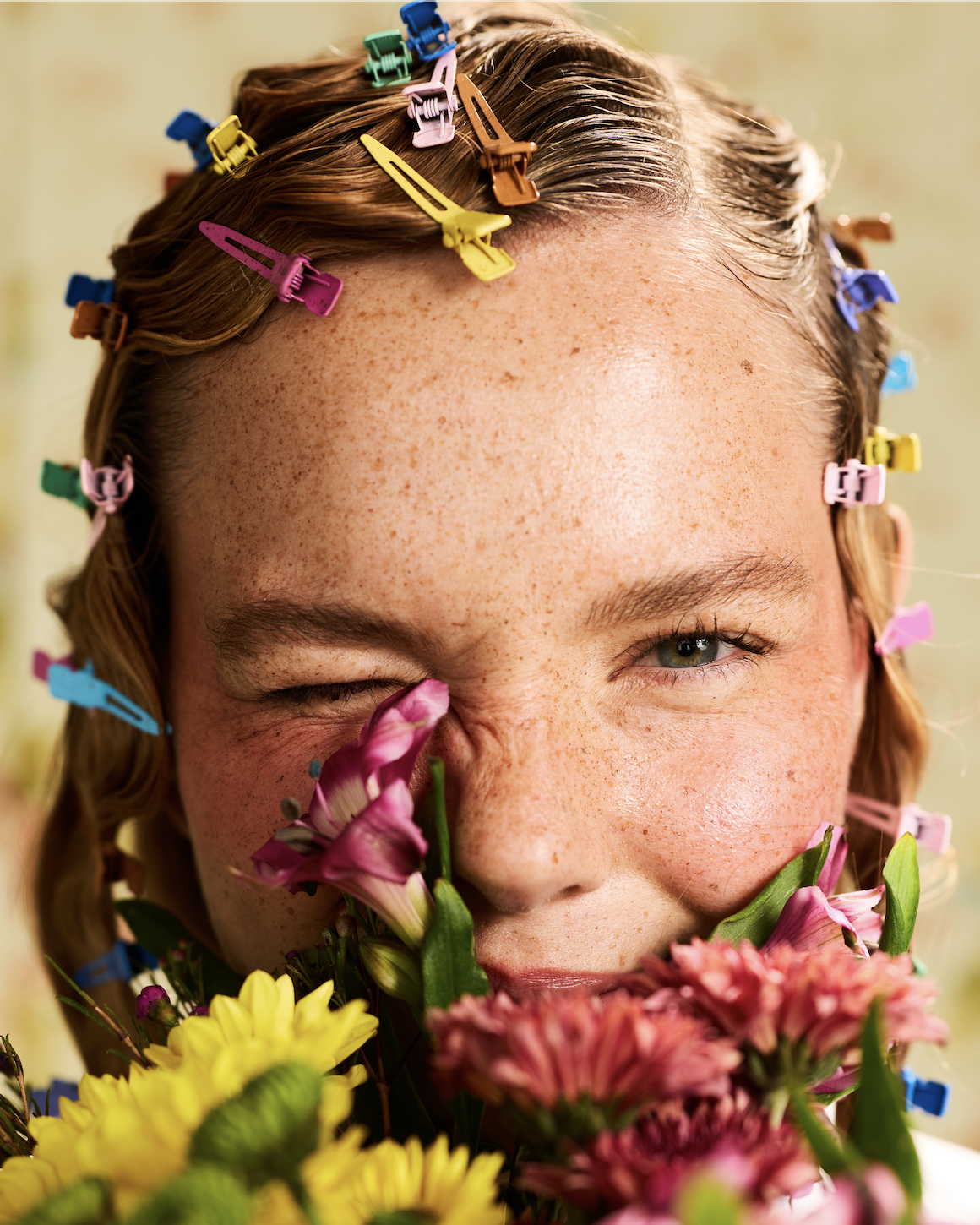 Girl with colorful hair clips, holding pink and yellow flowers near her face, winking and smiling.