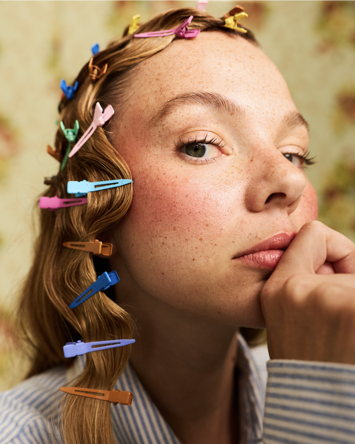 Close-up of a woman with red hair, blue eyes, and freckles, partially covered by her hand, with her hair styled and secured with colorful hair clips.