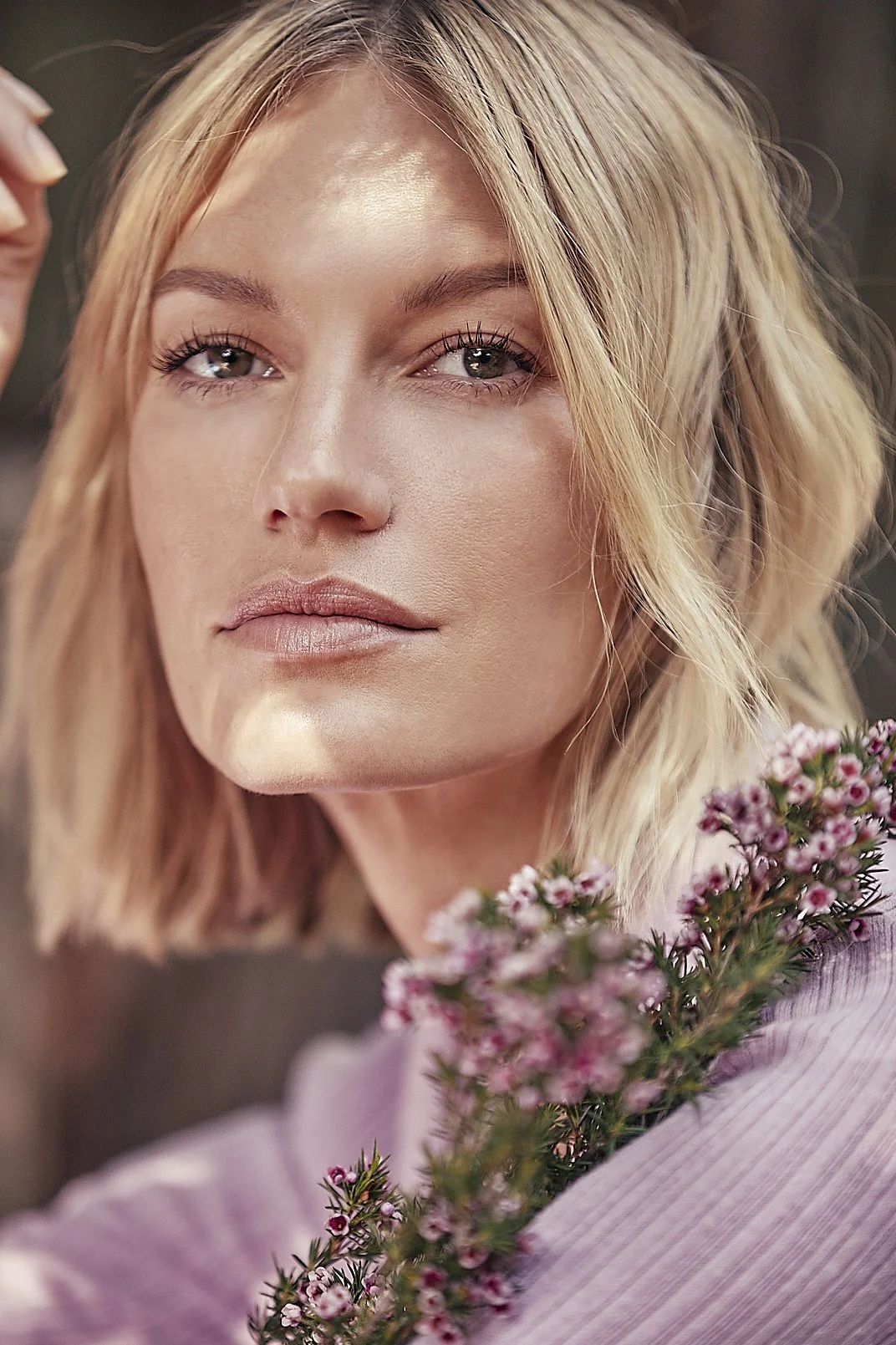Close-up portrait of a woman with blonde hair, blue eyes, and natural makeup holding pink flowers.