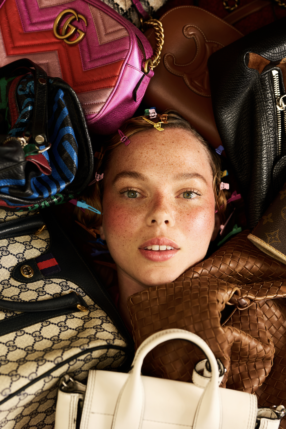 Close-up of a young woman with freckles and curly hair, surrounded by colorful designer handbags and accessories.