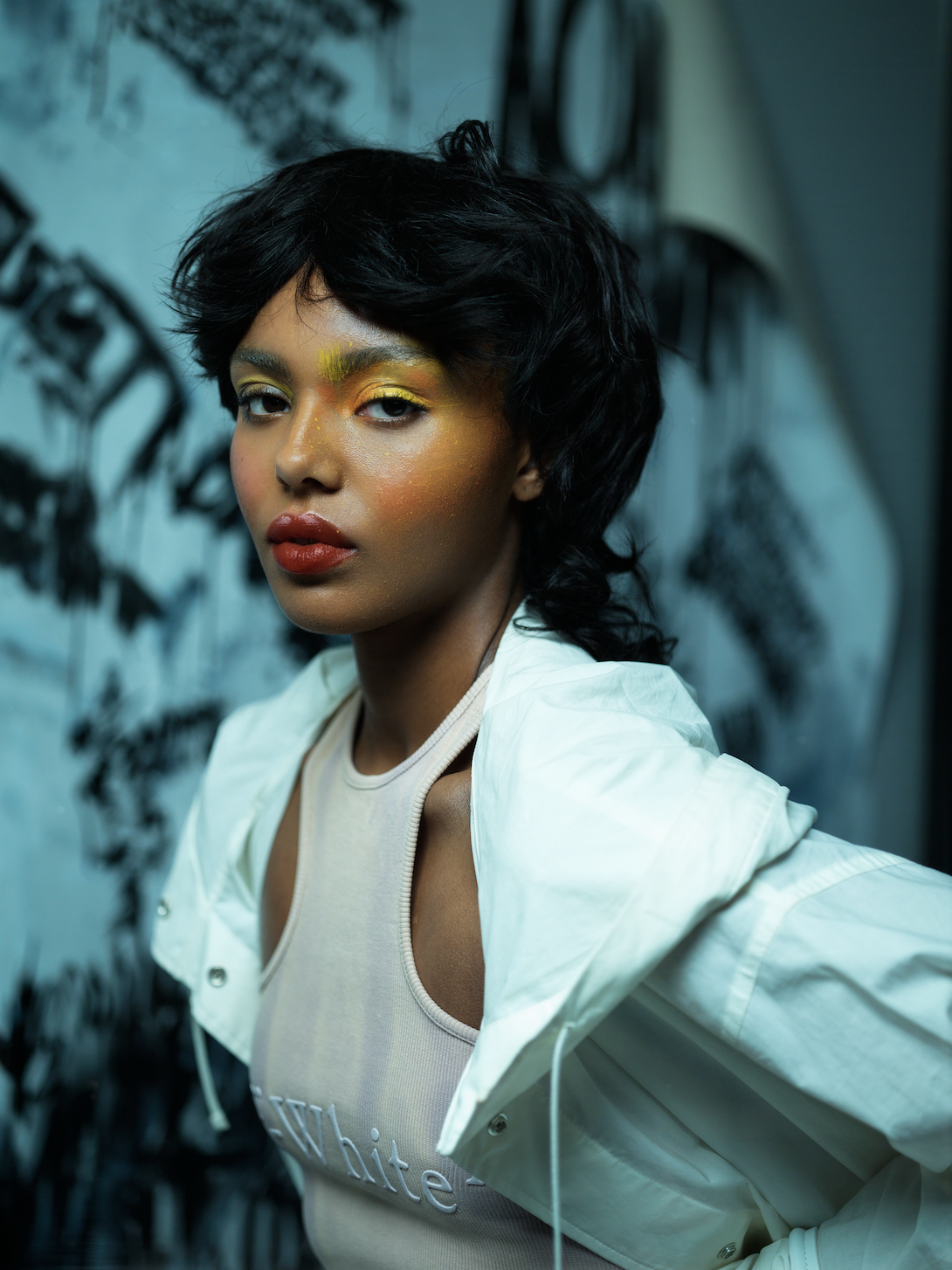 A young woman with short, black, wavy hair and vibrant gold and yellow makeup on her face, wearing a white tank top with 'white' written on it and a white windbreaker jacket, stands in front of a mural with black abstract shapes on a blue background.