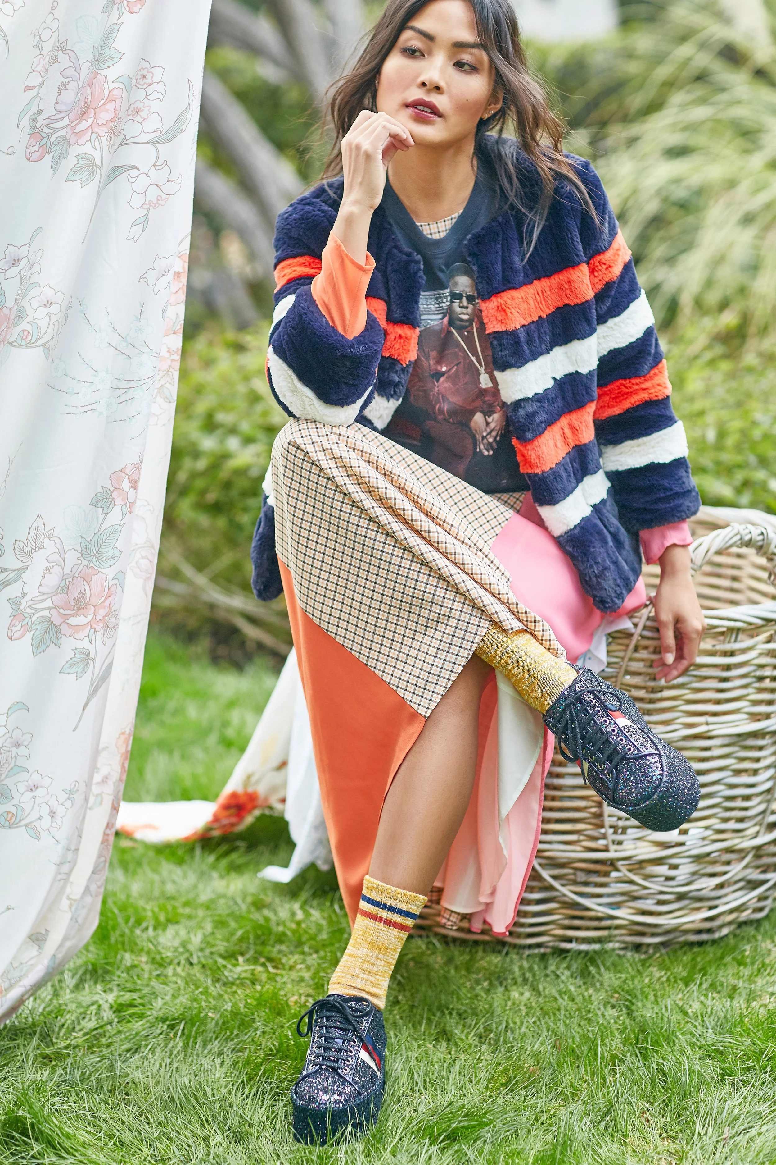 Young woman outdoors with one leg raised, wearing a colorful striped plush jacket, patterned pants, and glittery sneakers, sitting next to wicker chair and floral curtain, greenery in background.